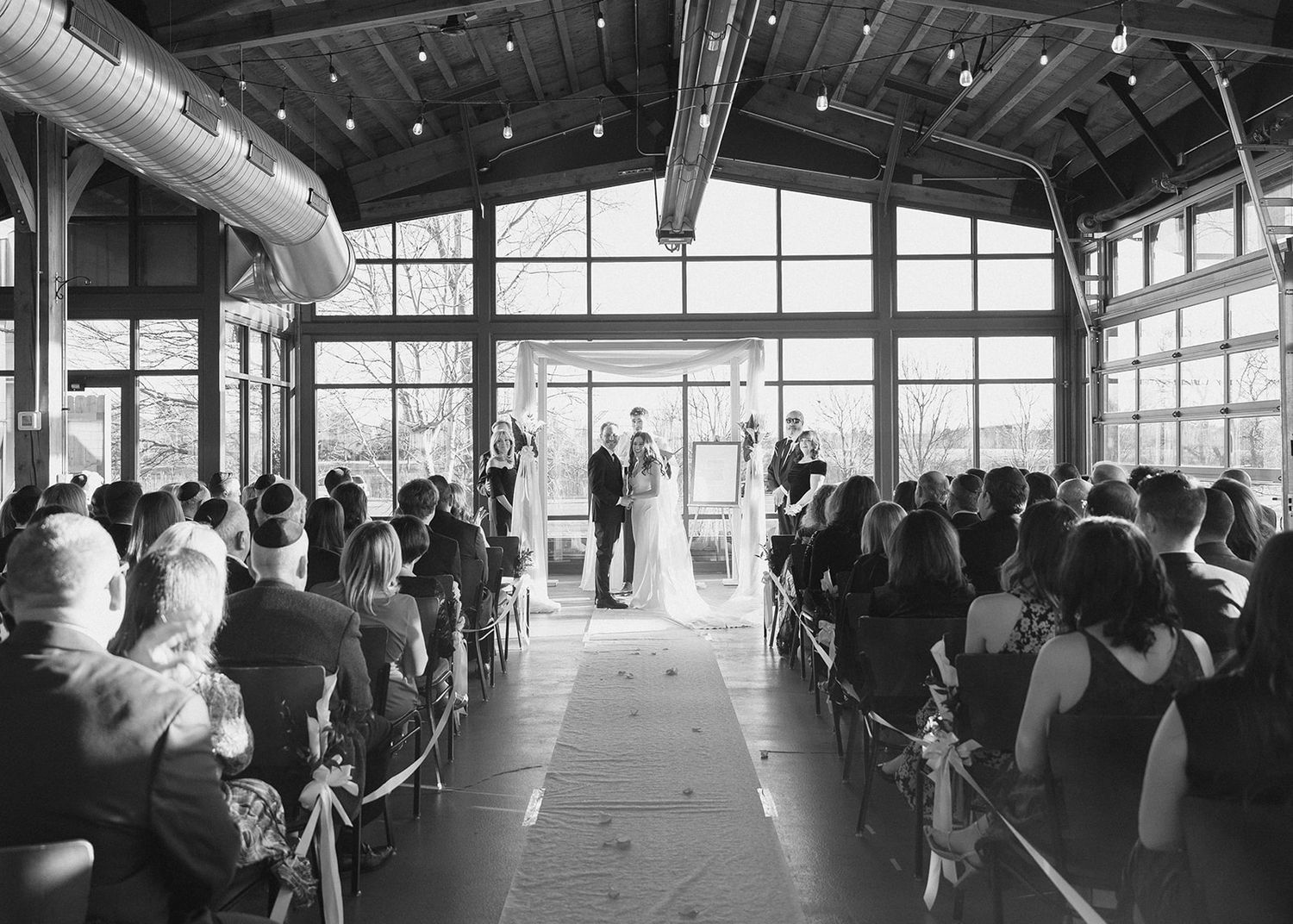 Black and white photo of a rustic indoor wedding ceremony in a glass-walled venue with seated guests.