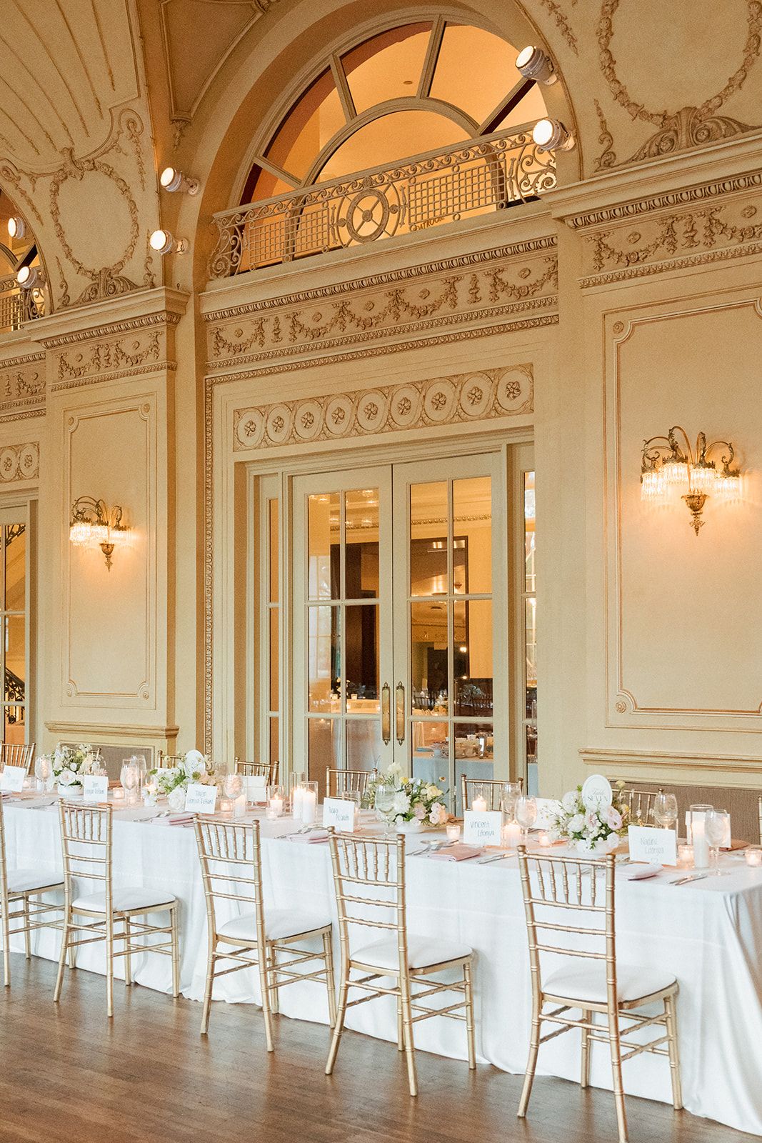 Elegant wedding reception hall with gold chiavari chairs, white linen tables, ornate moldings, and warm candlelight in a grand ballroom.