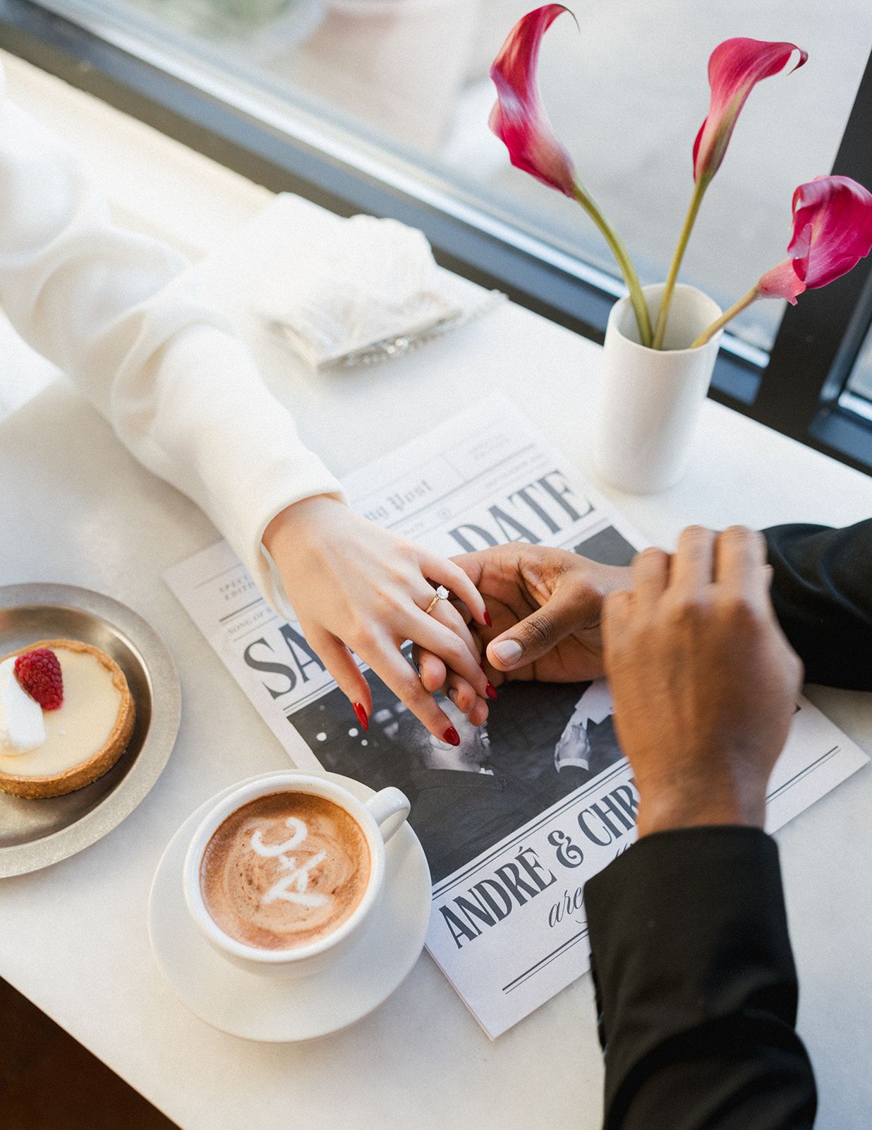 Overhead view of couple holding hands at cafe table with save the date card, coffee, and pink flowers.