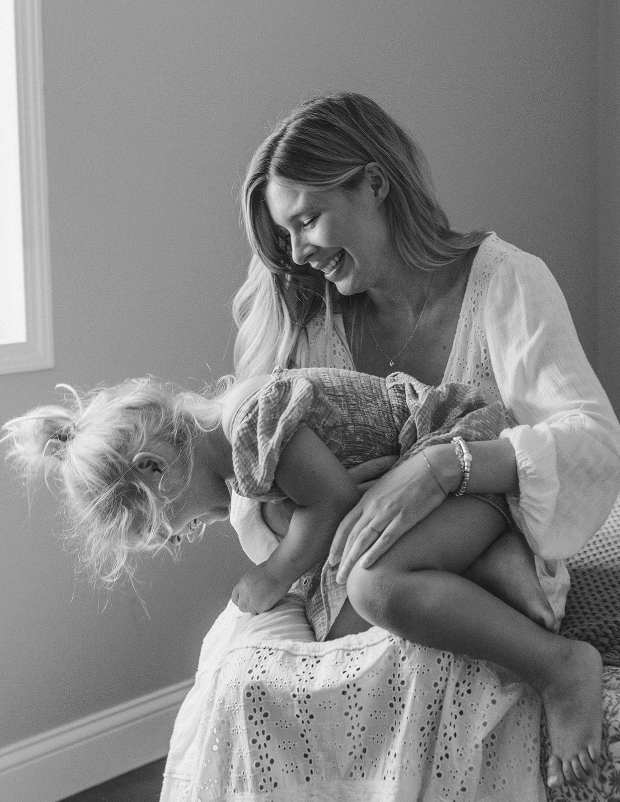 Black and white photo of a laughing mother holding her young daughter on her lap in a bright room.