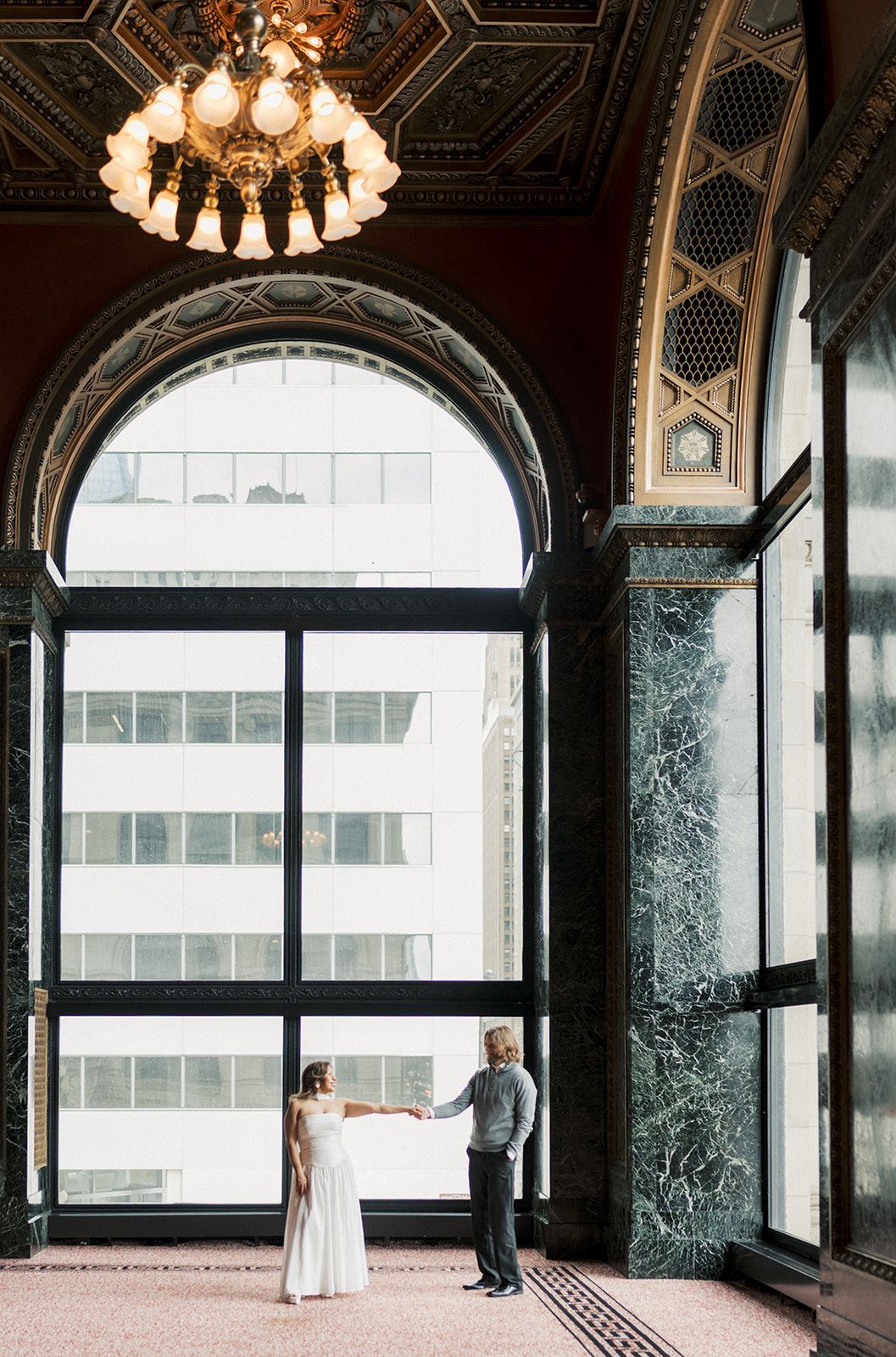 Couple holds hands in elegant marble-columned hall with arched windows and ornate chandelier.