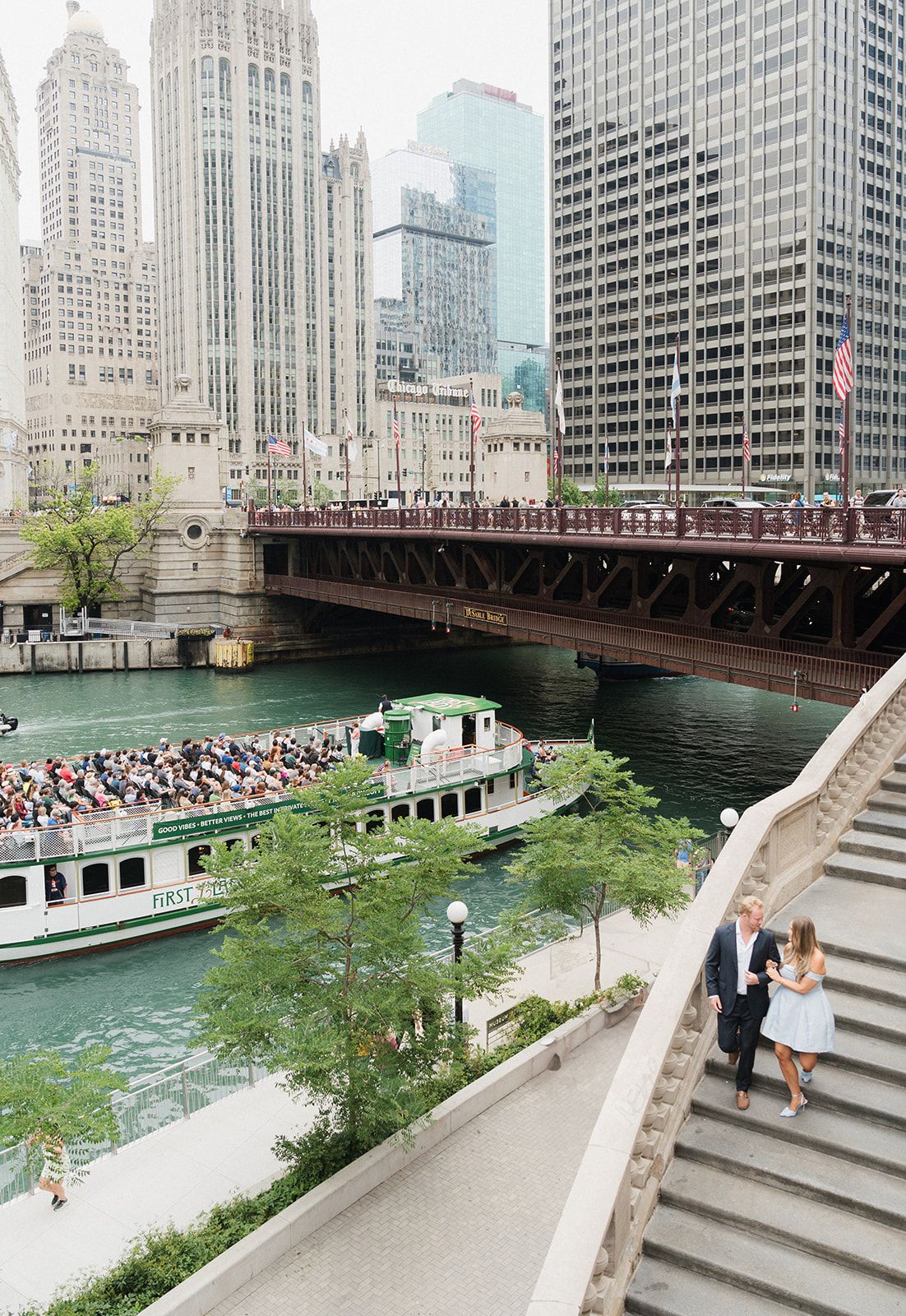 Couple walking up riverside stairs with Chicago River, bridge, and city skyline in background.