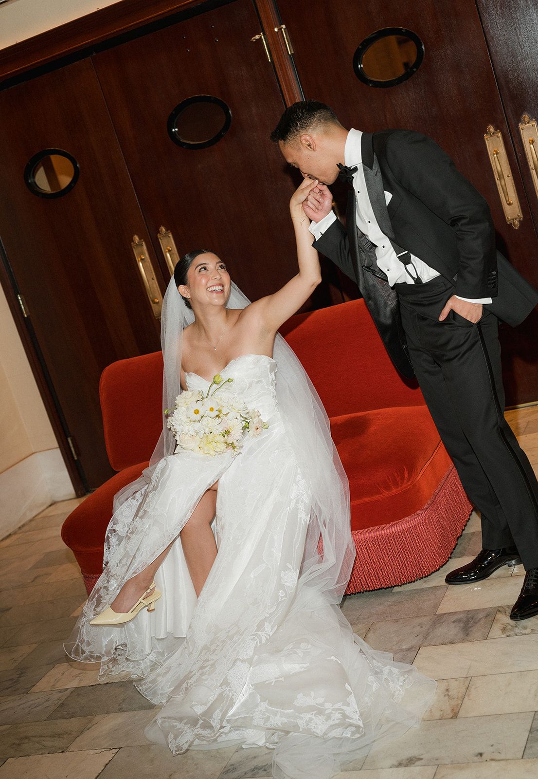 Bride in strapless white gown laughs as groom in black tuxedo kisses her hand beside a red velvet sofa.