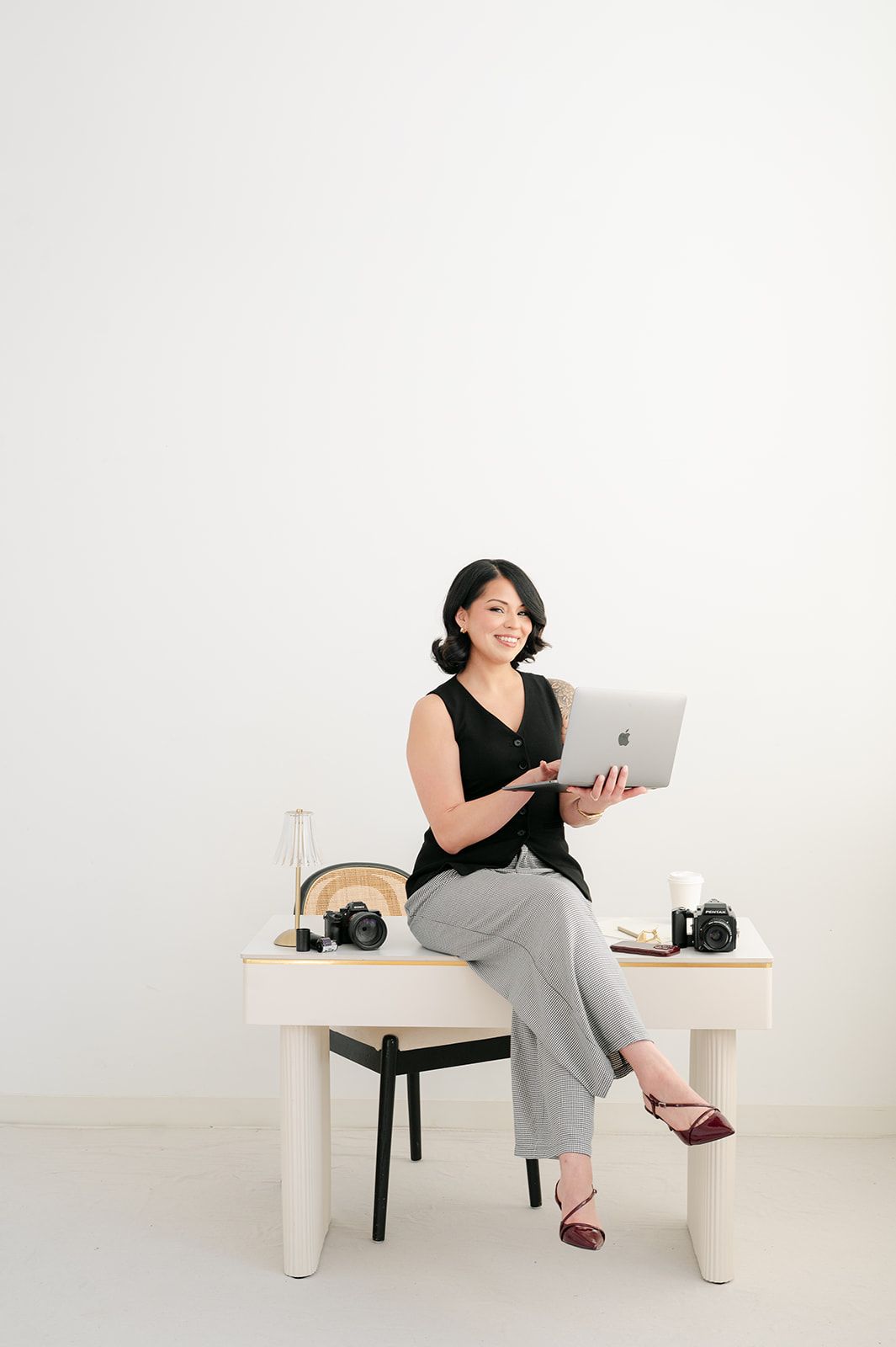 Professional female photographer sits on desk holding laptop surrounded by cameras in bright studio.