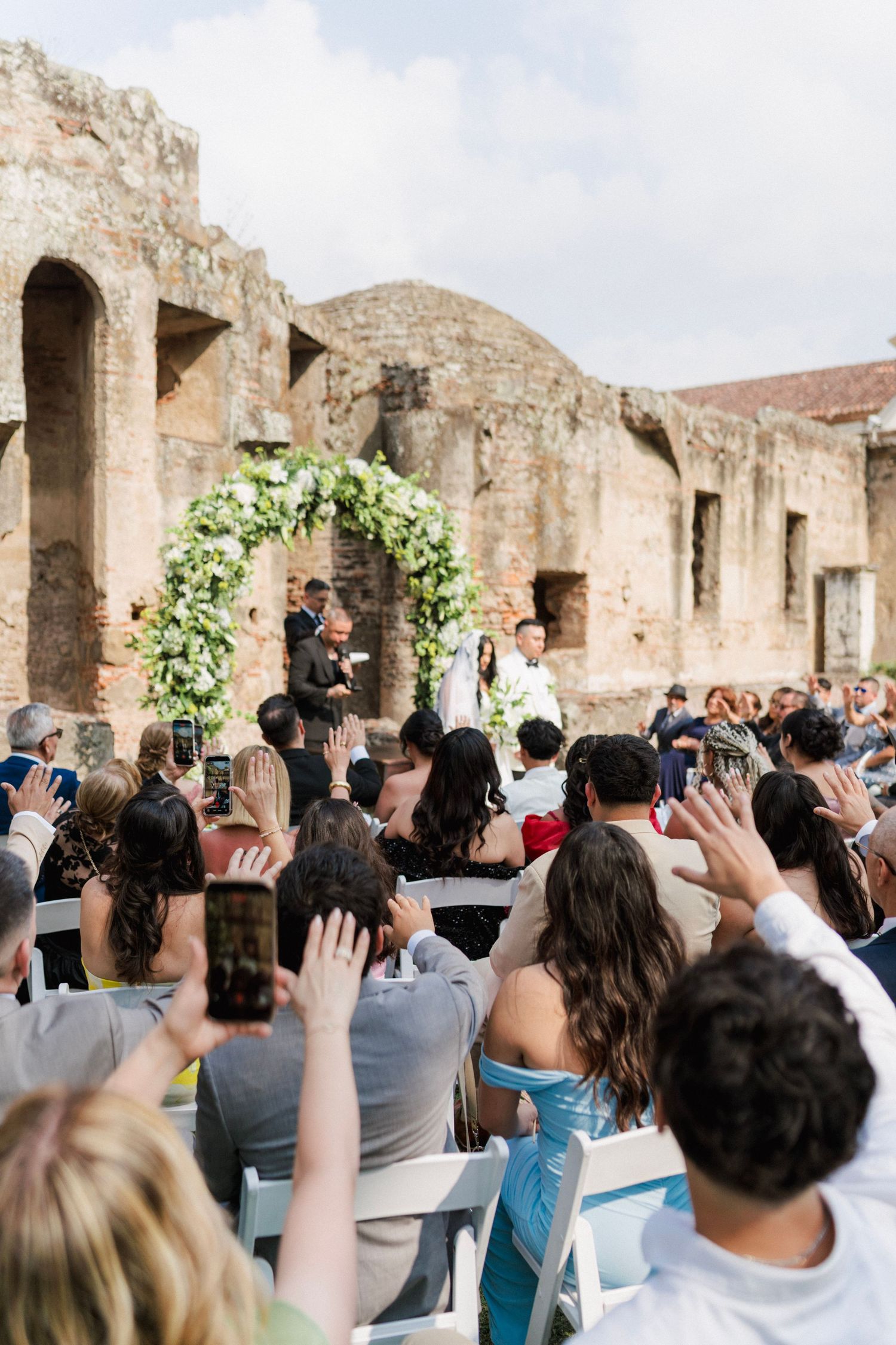 Guests applaud at an outdoor wedding ceremony amid ancient stone ruins, with couple at floral arch altar.