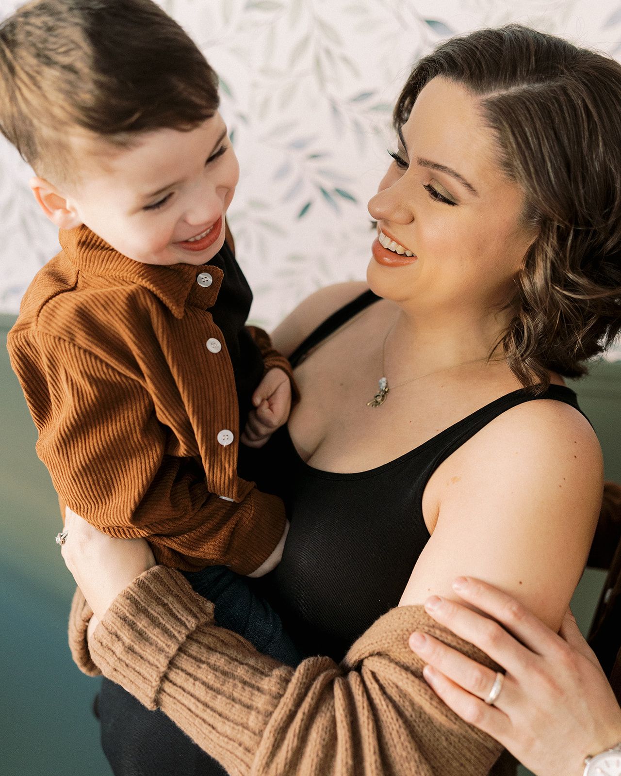 A smiling mother holds her happy toddler son dressed in a brown corduroy shirt in a warm embrace.