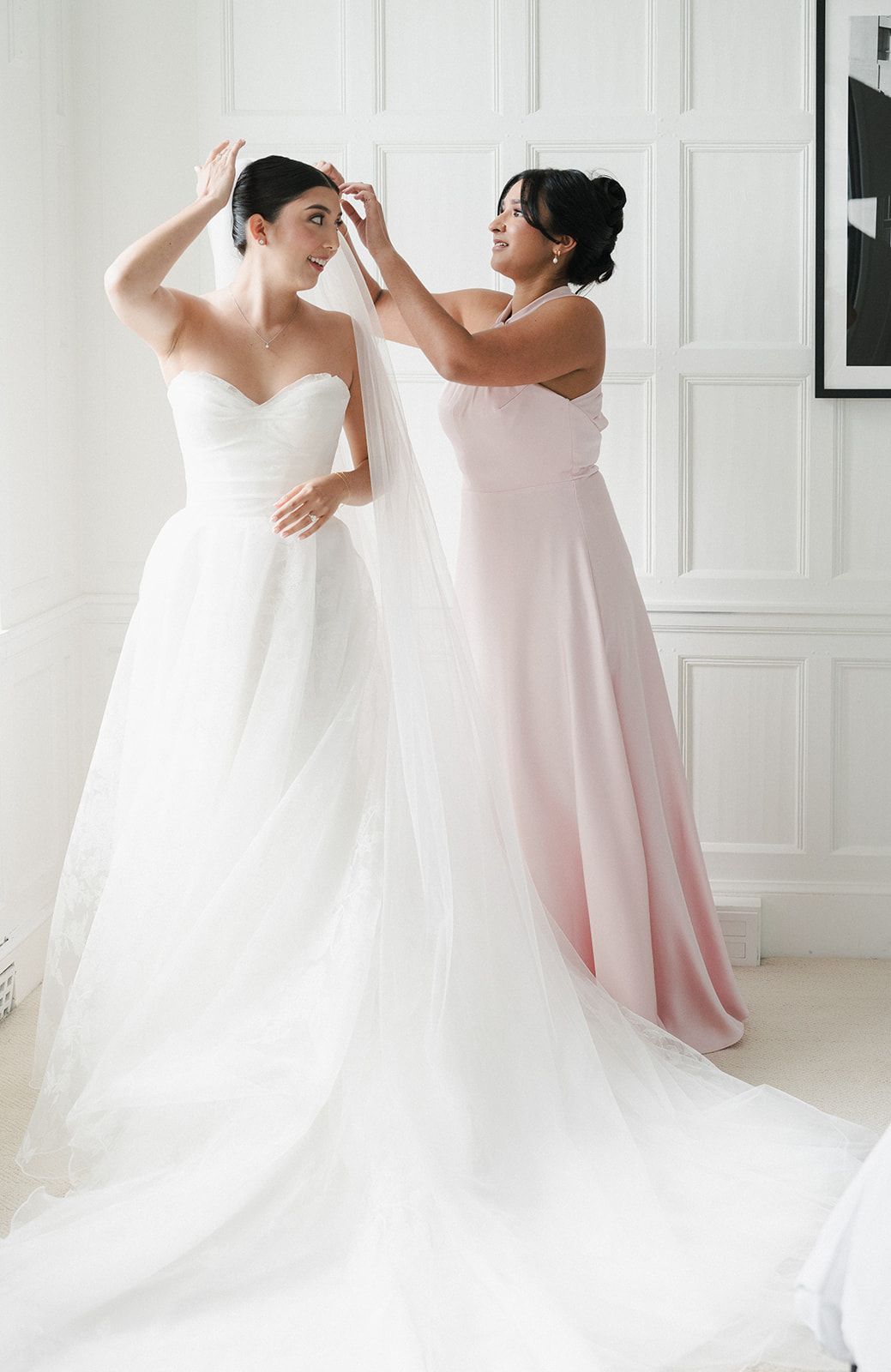 Bride in white strapless gown gets veil adjusted by bridesmaid in blush pink dress in bright white room.