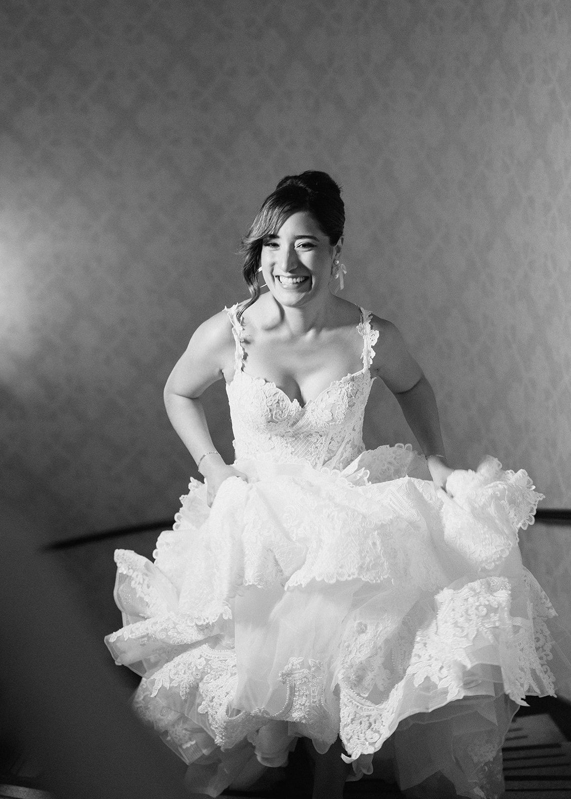 Joyful bride in lace ball gown laughs while adjusting her dress in black and white photo.