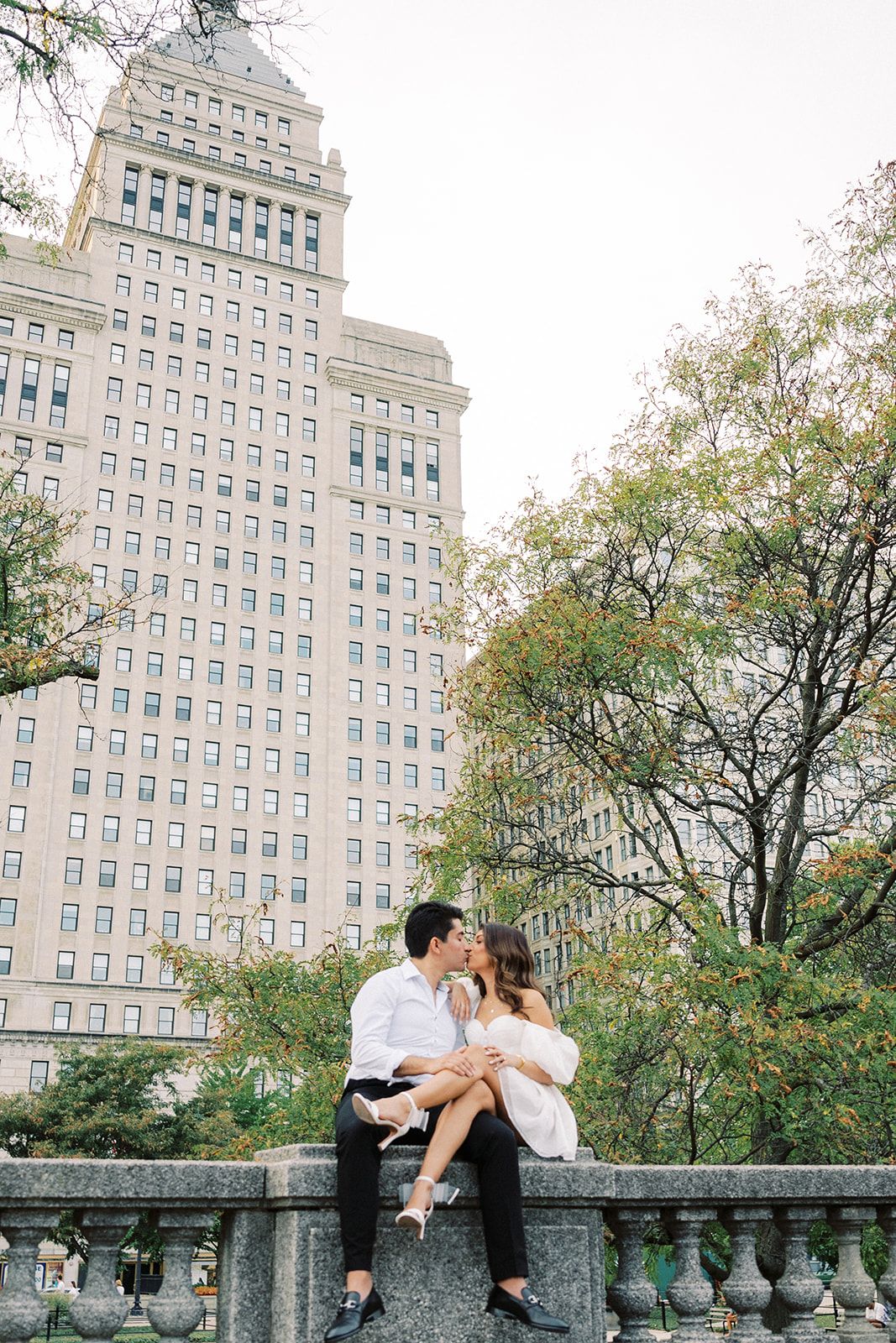 A couple shares a romantic kiss while sitting on a stone balustrade with a tall city building and green trees behind them.