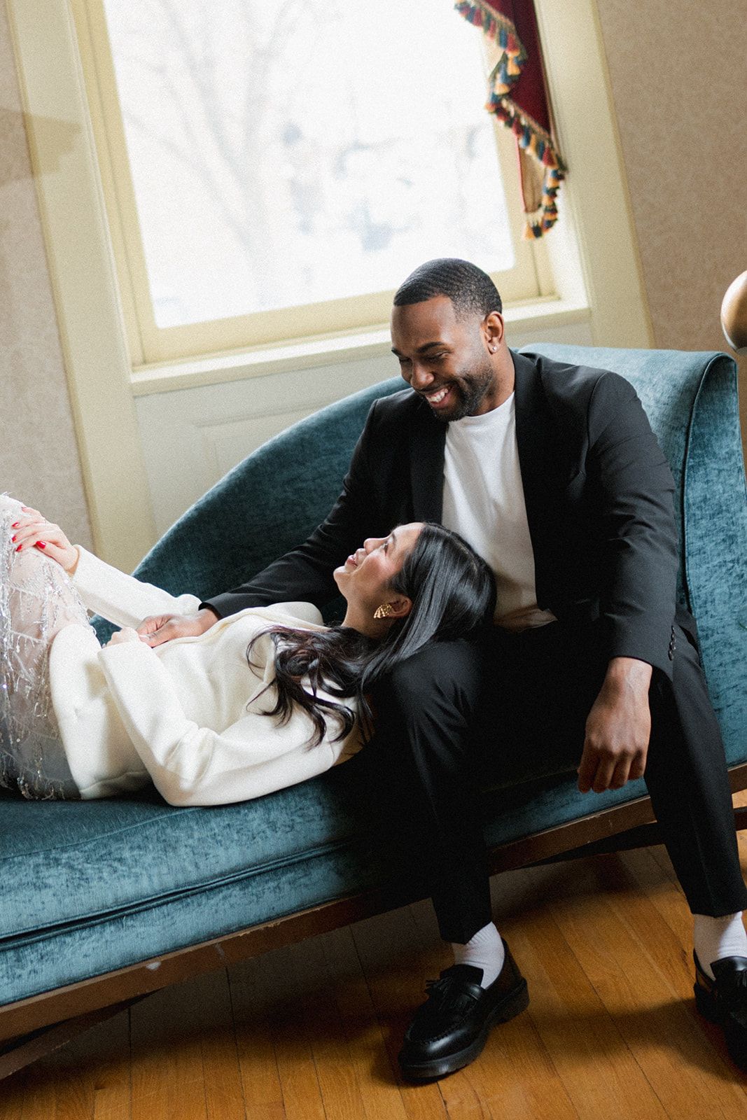 Smiling couple relaxing on a teal velvet chaise lounge, man in black suit, woman in white outfit.