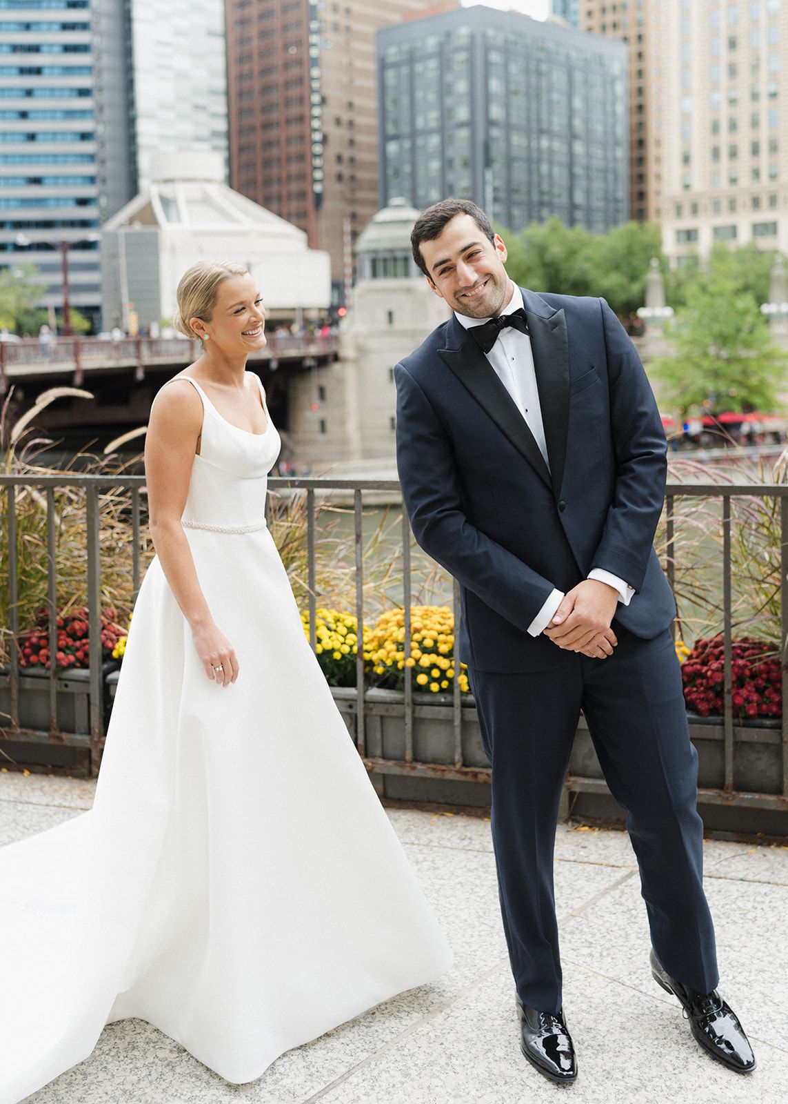 Smiling bride and groom in navy tuxedo posing on bridge with Chicago skyline backdrop.