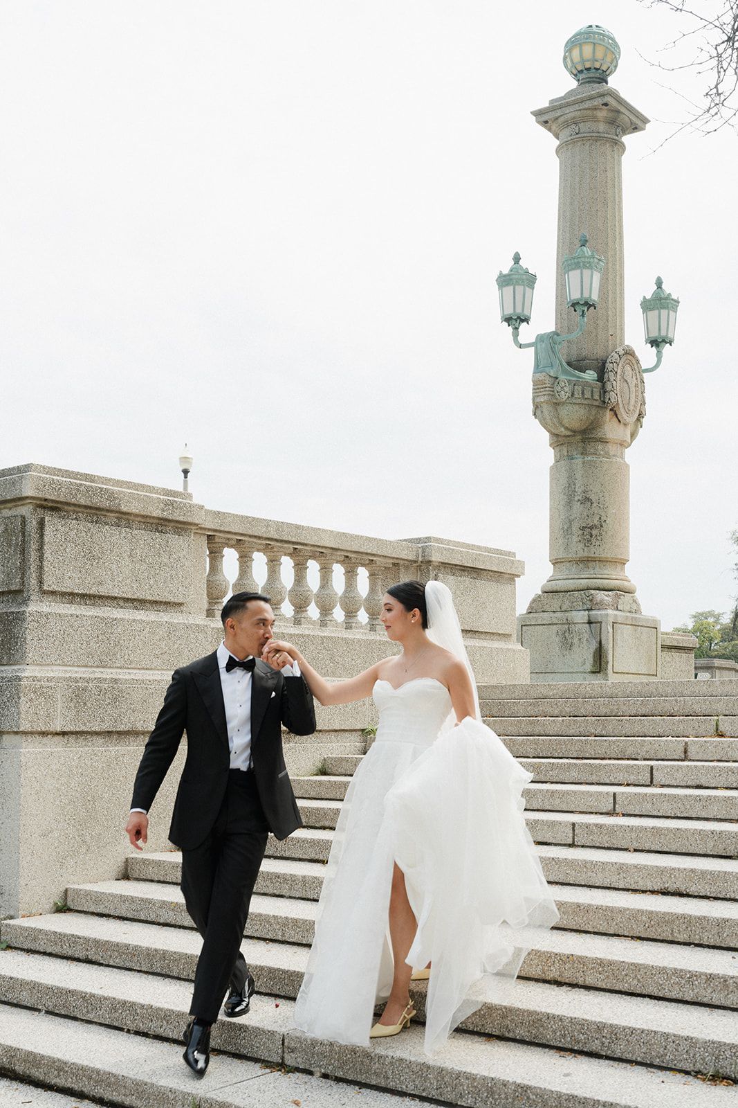 Bride and groom descending grand stone steps near an ornate lamppost on their wedding day.