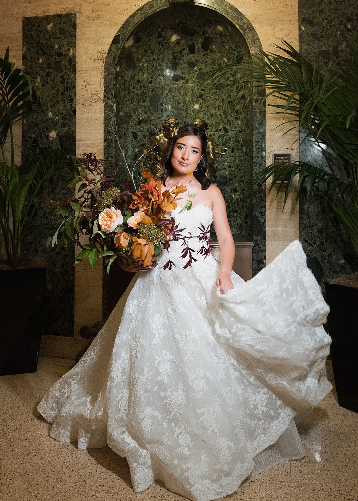 Bride in lace ballgown holds autumn bouquet of orange and burgundy blooms before marble arch backdrop.