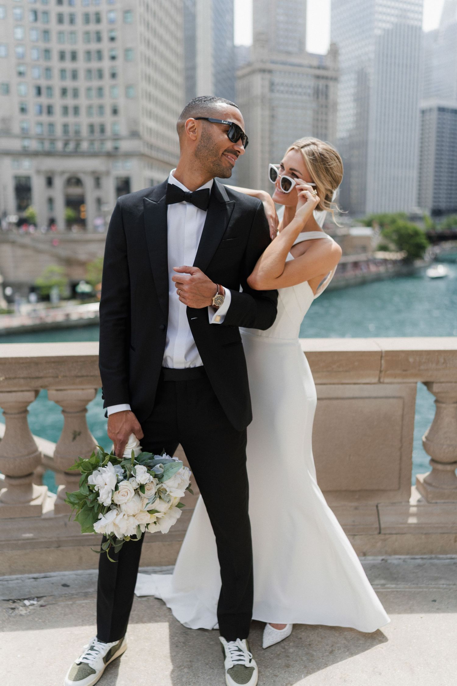 Bride and groom in sunglasses pose on a Chicago bridge, she in a white gown, he in a black tuxedo with sneakers.