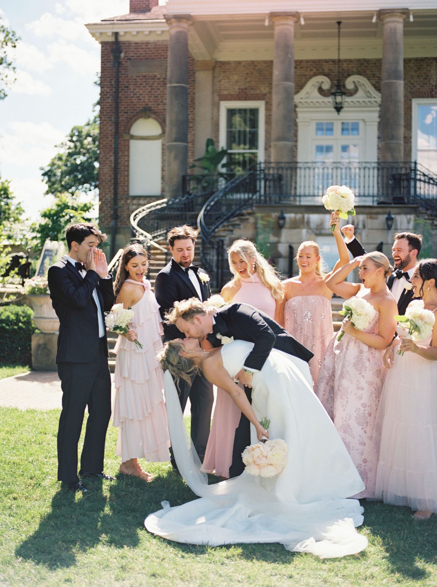 Bride and groom share a dip kiss surrounded by wedding party in pink gowns and tuxedos outside a brick mansion.