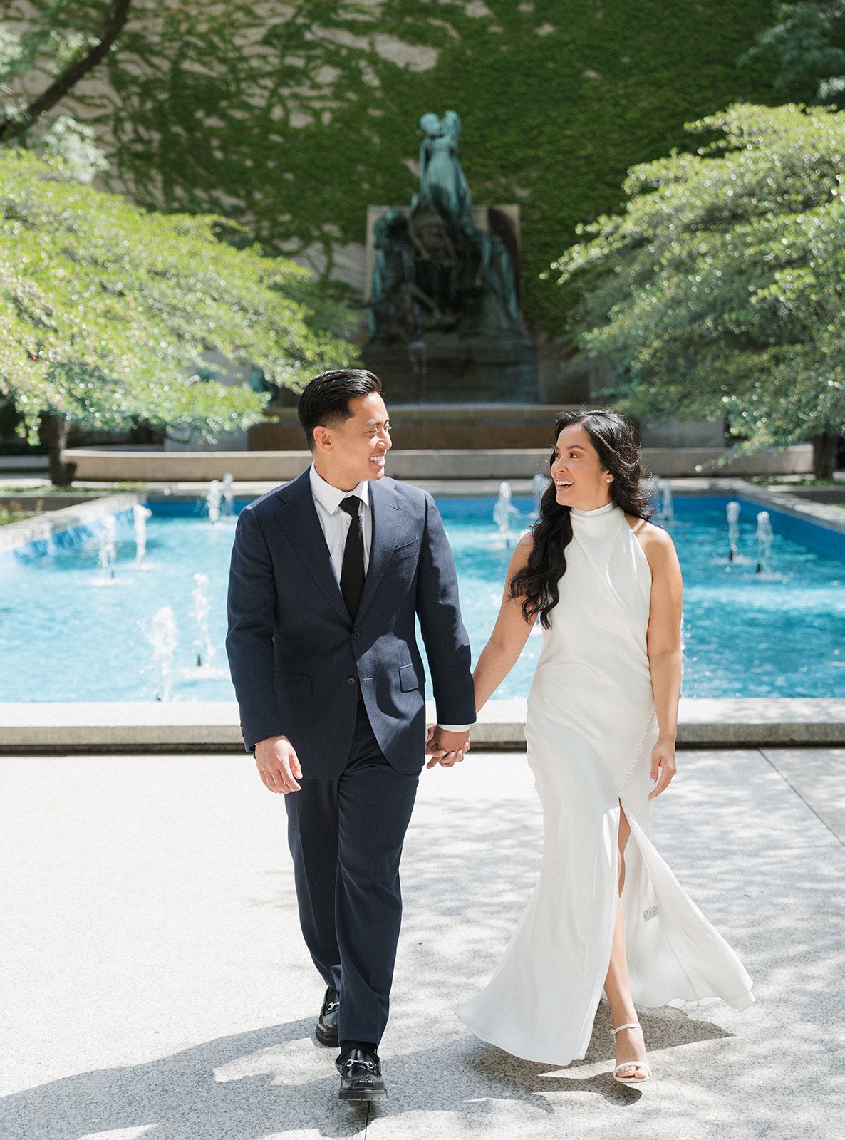 Couple in wedding attire holding hands by a fountain, man in navy suit, woman in white gown, lush green trees backdrop.
