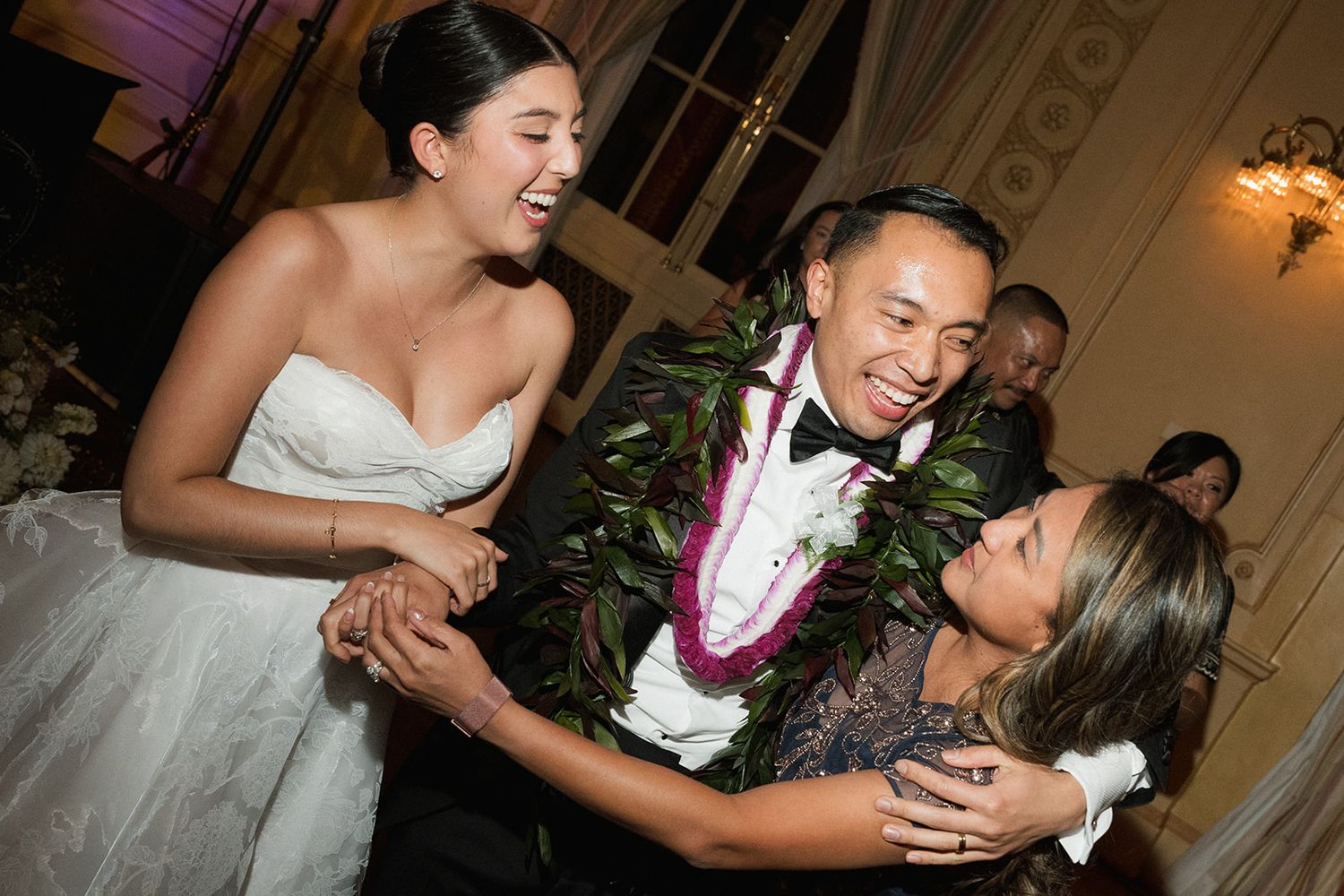 Bride and groom laughing with guest at Hawaiian wedding reception, groom wearing traditional lei garlands.