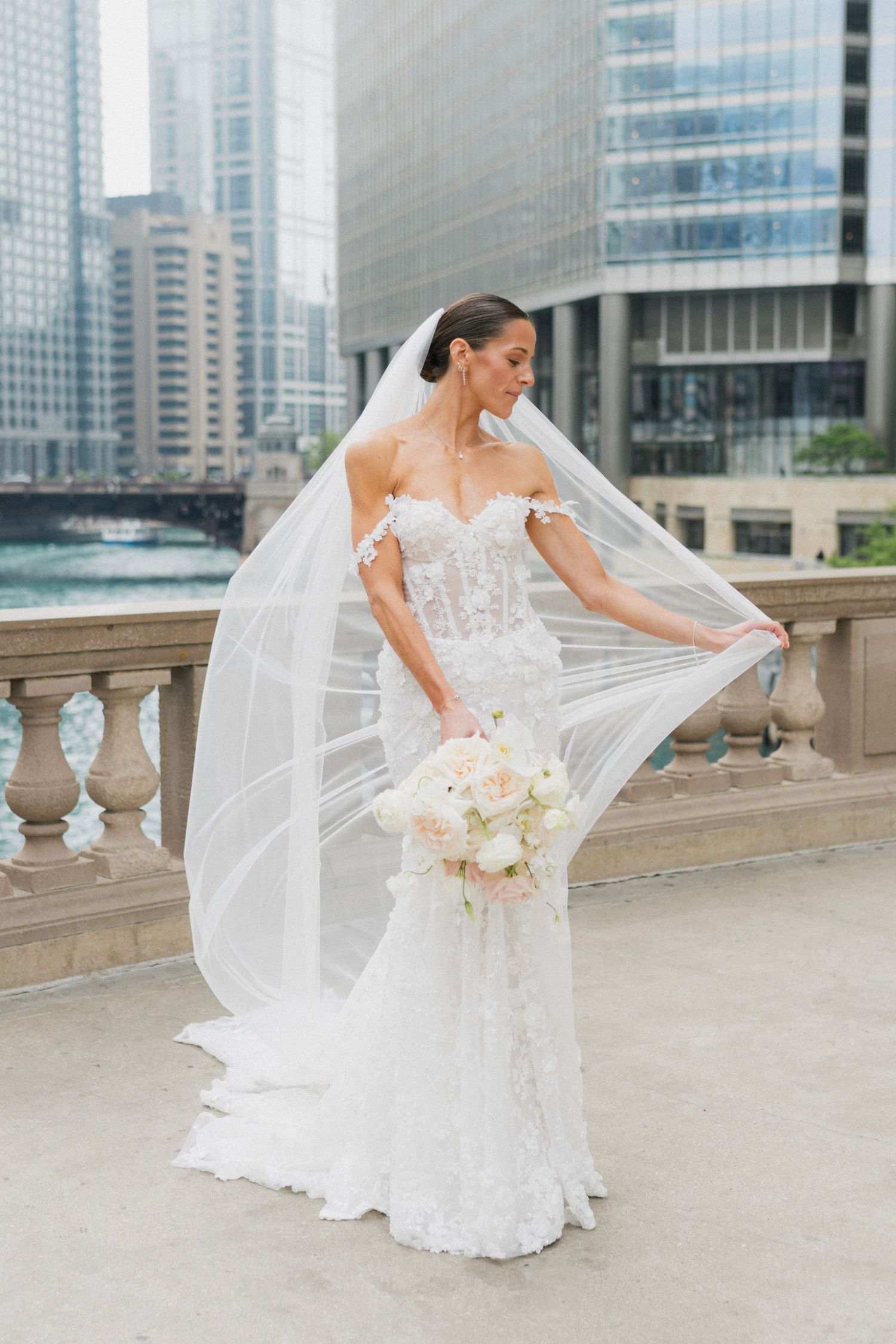 Bride in lace off-shoulder gown holds bouquet, veil flowing, posed on urban bridge with city skyline backdrop.