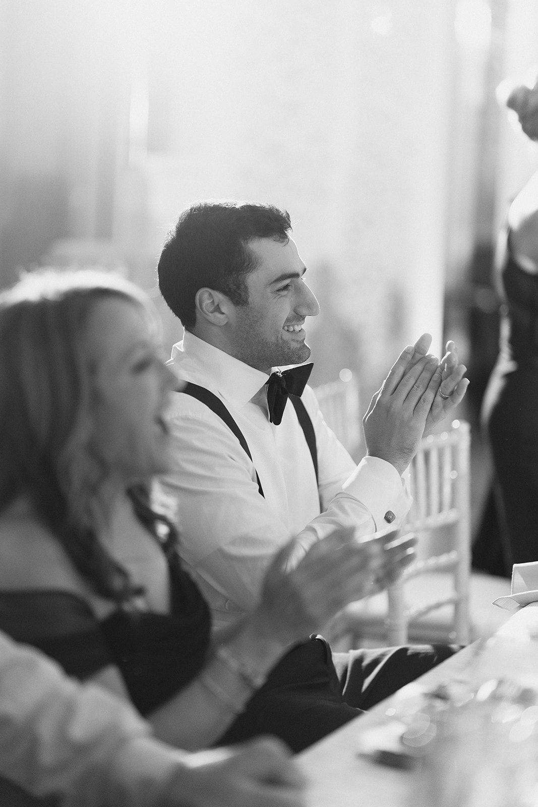 Black and white photo of a smiling groom in a bow tie clapping at a wedding reception.