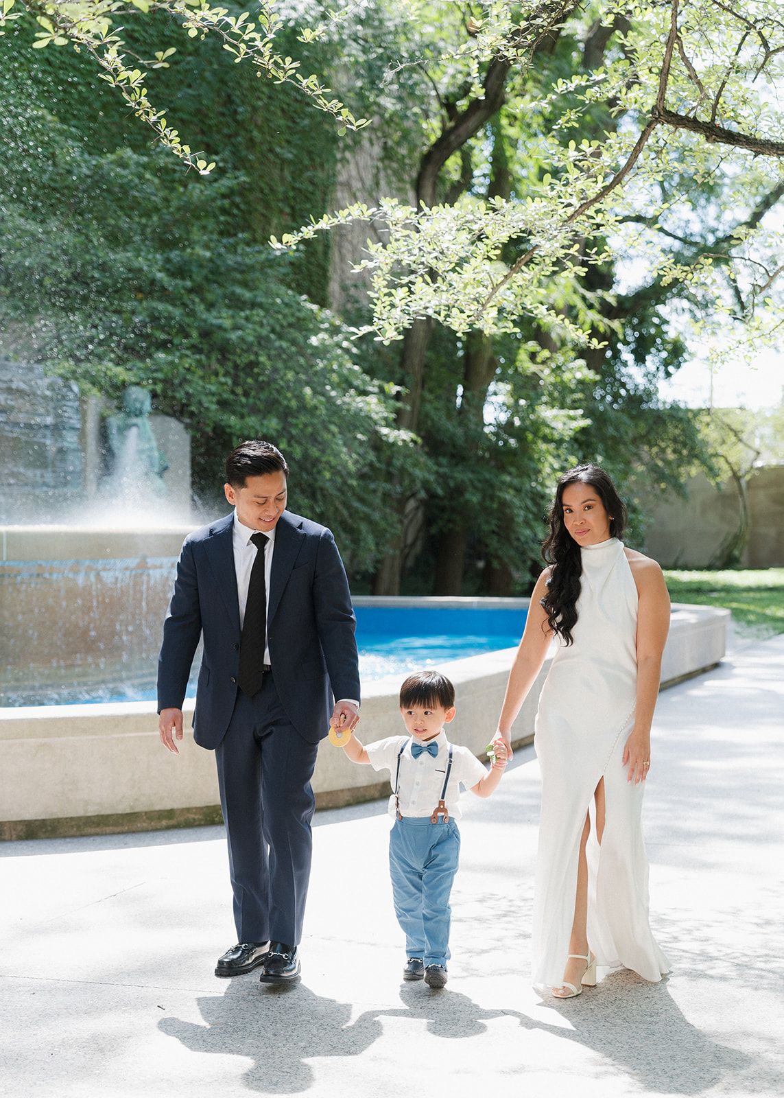 A bride and groom in elegant wedding attire walk hand-in-hand with their young son near a fountain.