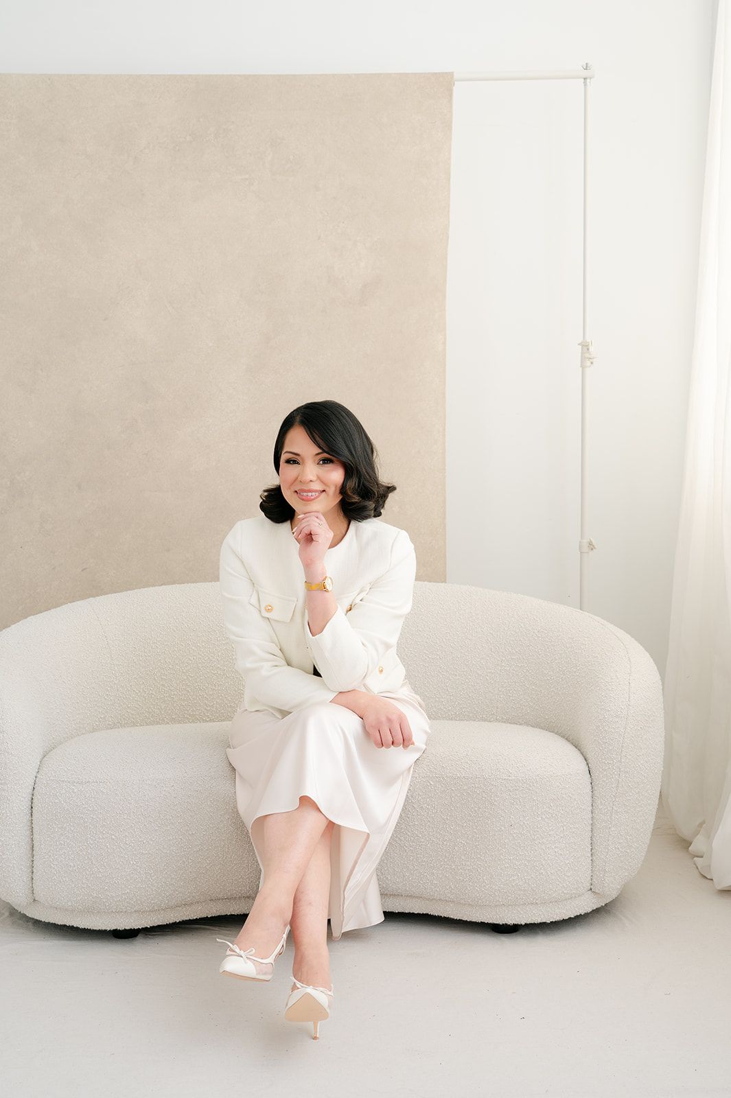 Elegant woman in white blazer and skirt sitting on a curved cream sofa in a bright room.