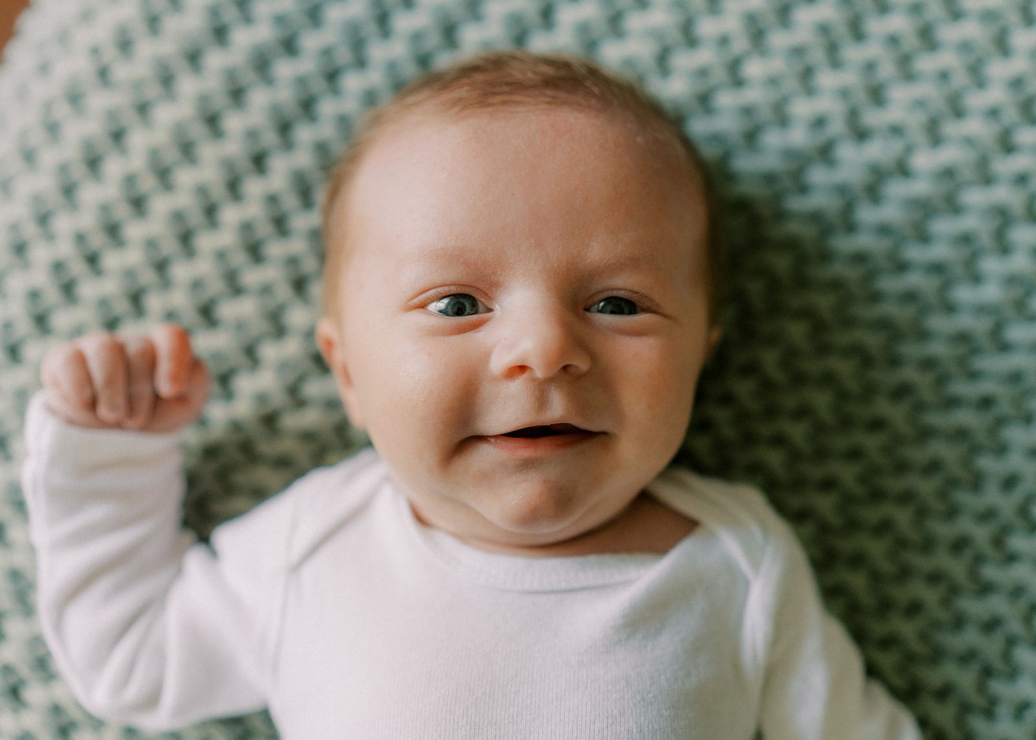 Smiling newborn baby in white onesie lying on green knit blanket with bright eyes and raised fist.