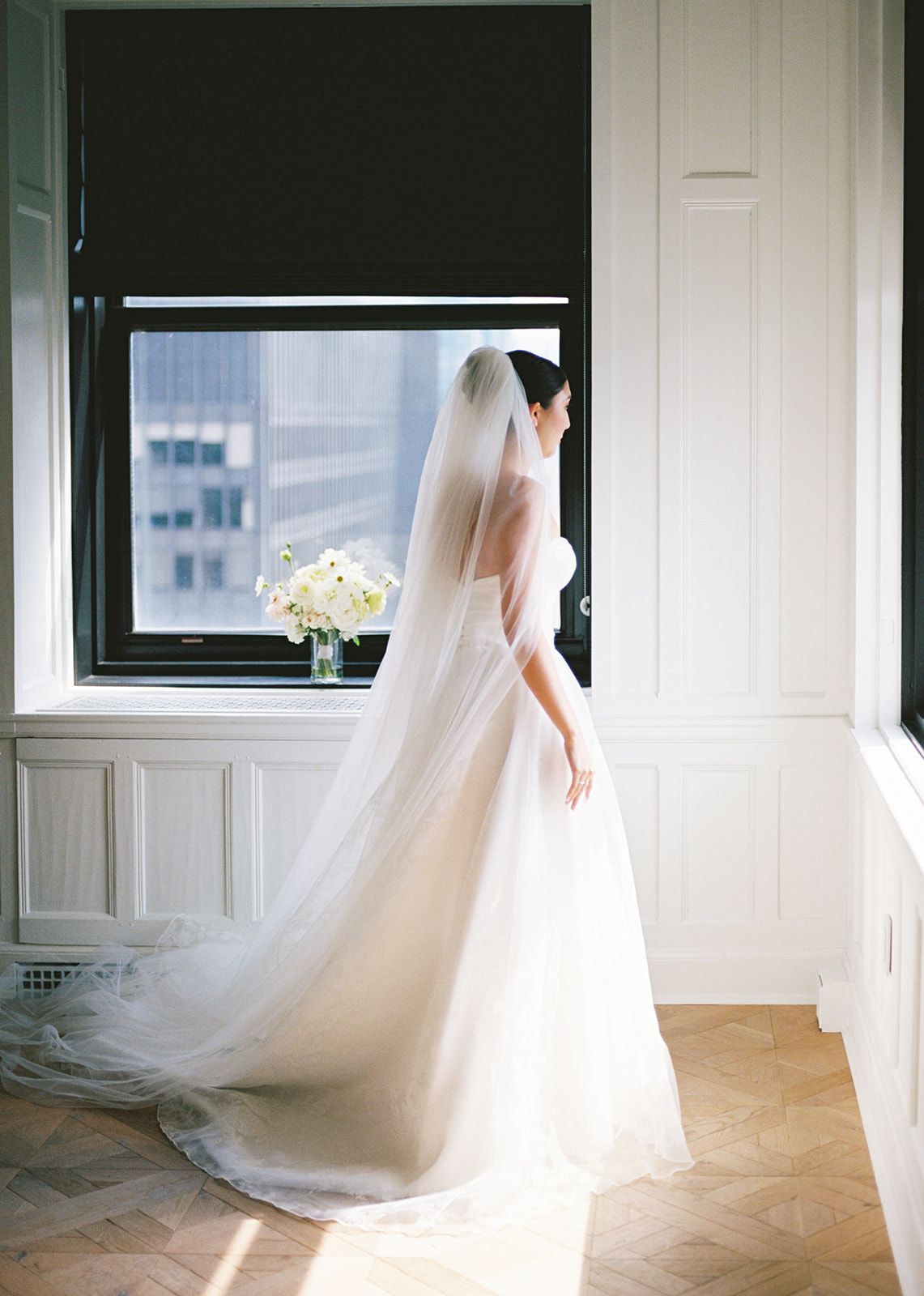 Bride in strapless ballgown with long cathedral veil stands by large window holding white floral bouquet indoors.