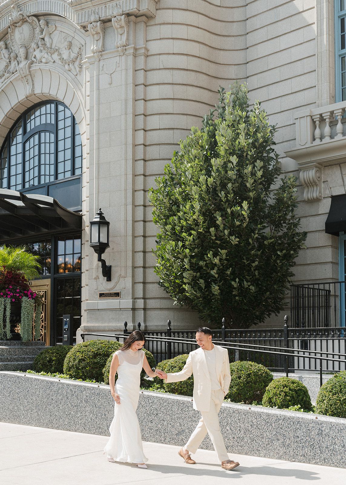 Bride in sleek white gown and groom in ivory suit walk hand-in-hand outside elegant beaux-arts building.