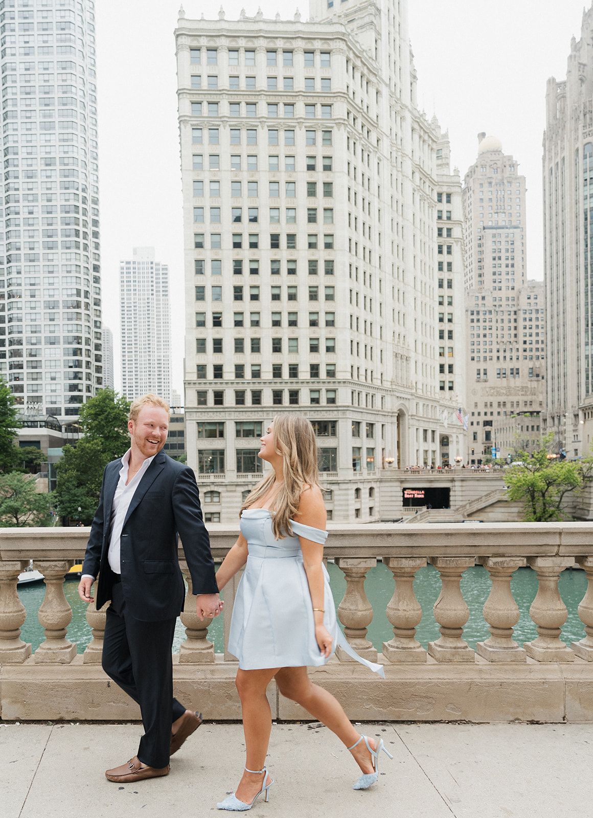 Couple holding hands walking on a Chicago bridge with the Wrigley Building skyline backdrop.
