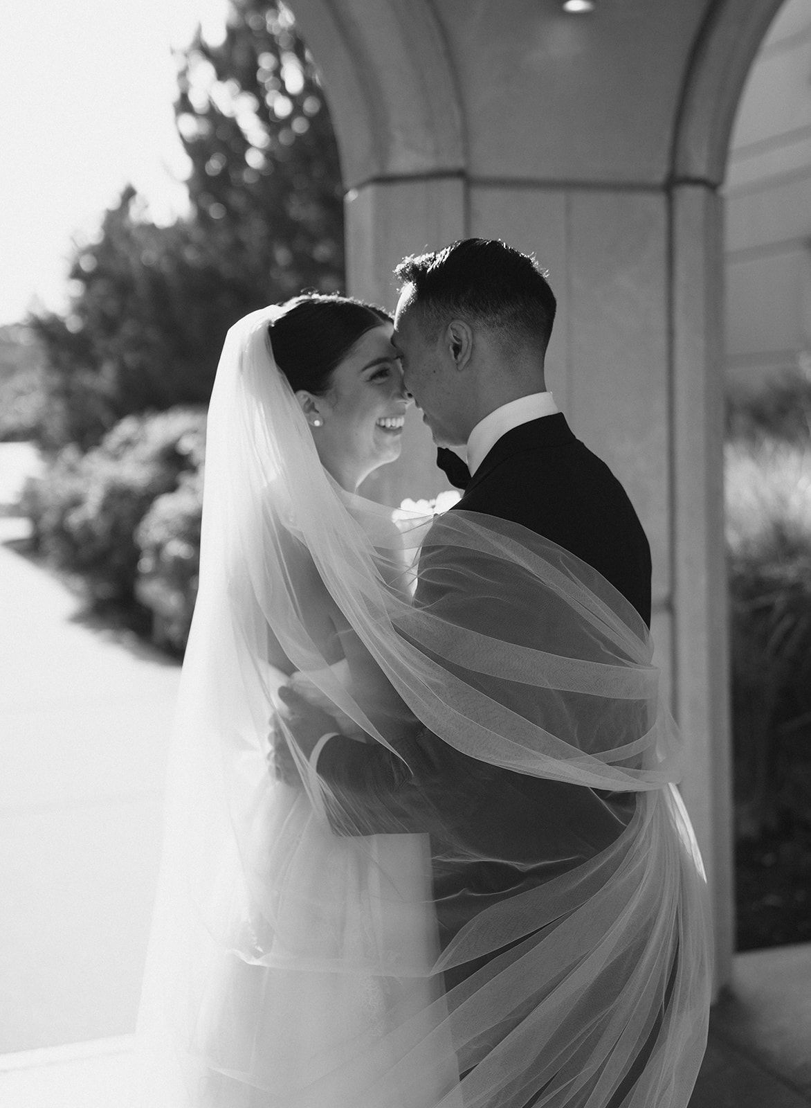 Black and white photo of bride and groom embracing, bride's veil flowing around them at outdoor wedding venue.