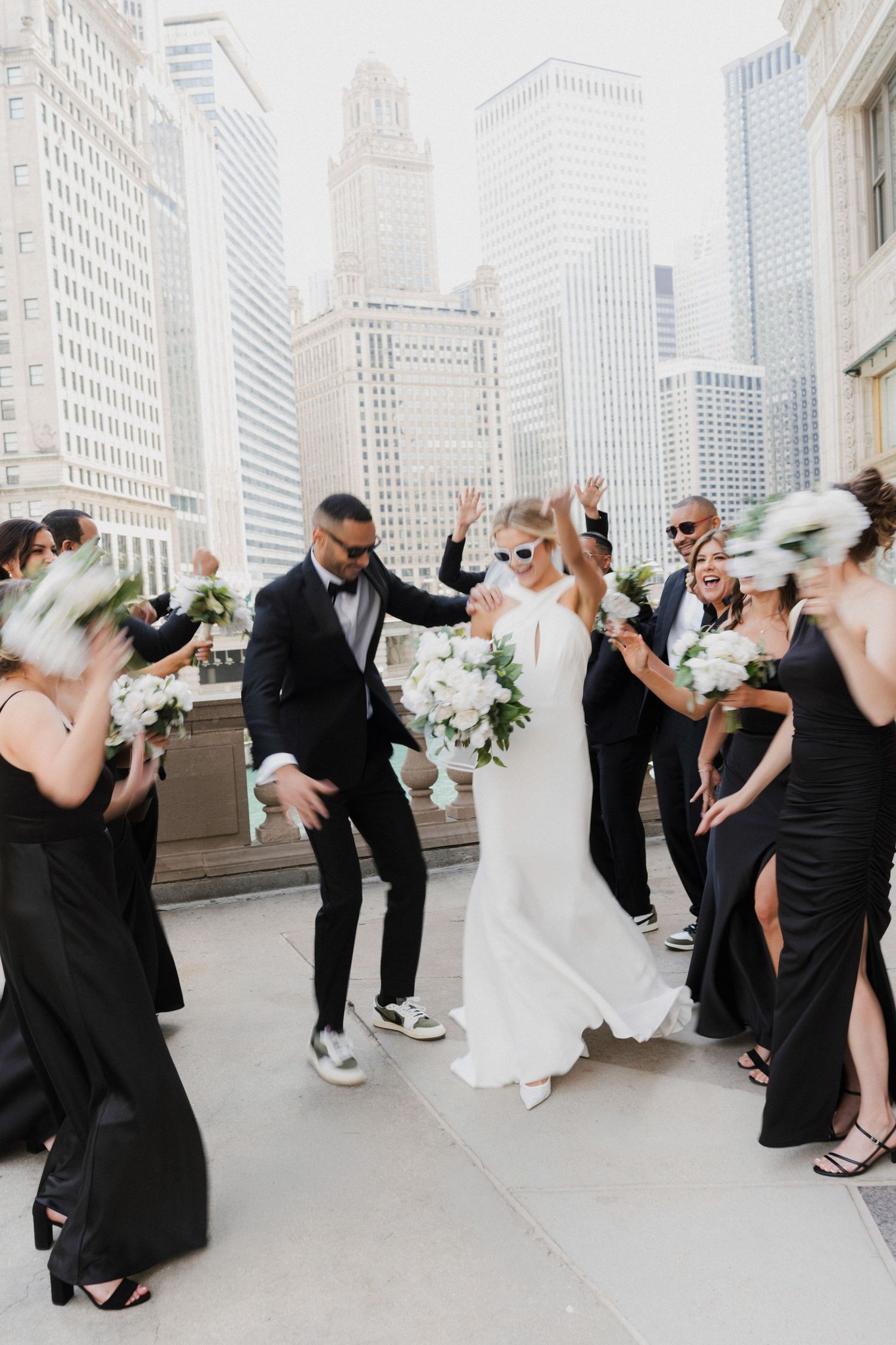 Bride and groom celebrate with wedding party in black attire on a rooftop with Chicago skyline backdrop.