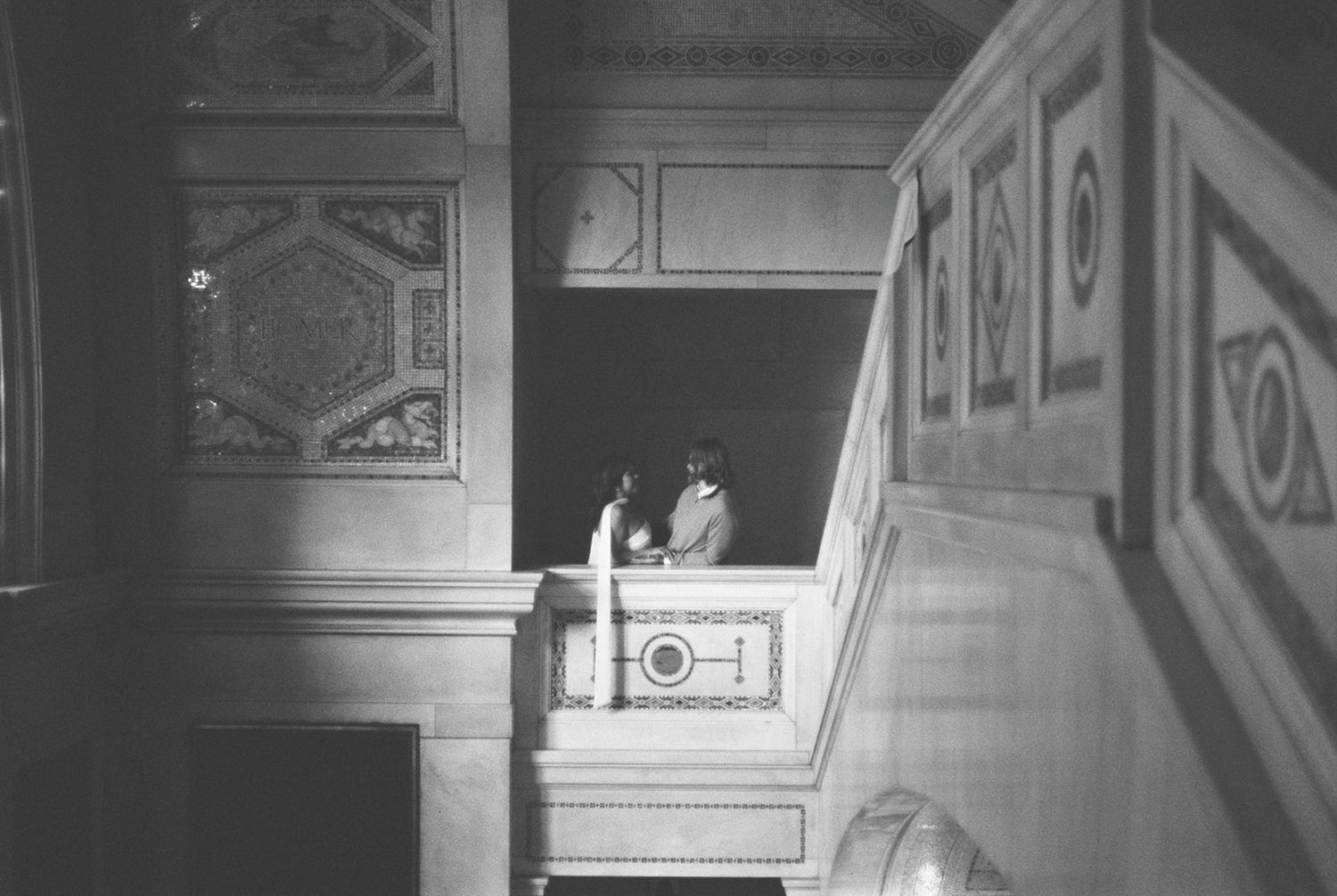 Black and white photo of two people standing on an ornate balcony inside a grand historic building.