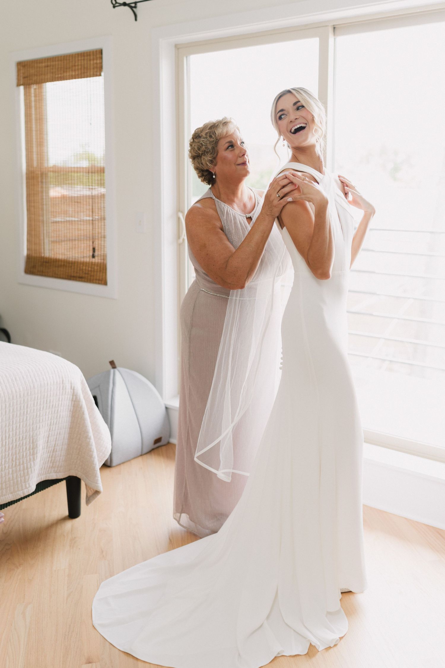 A bride laughing joyfully as an older woman helps her into her elegant white wedding gown in a bright room.