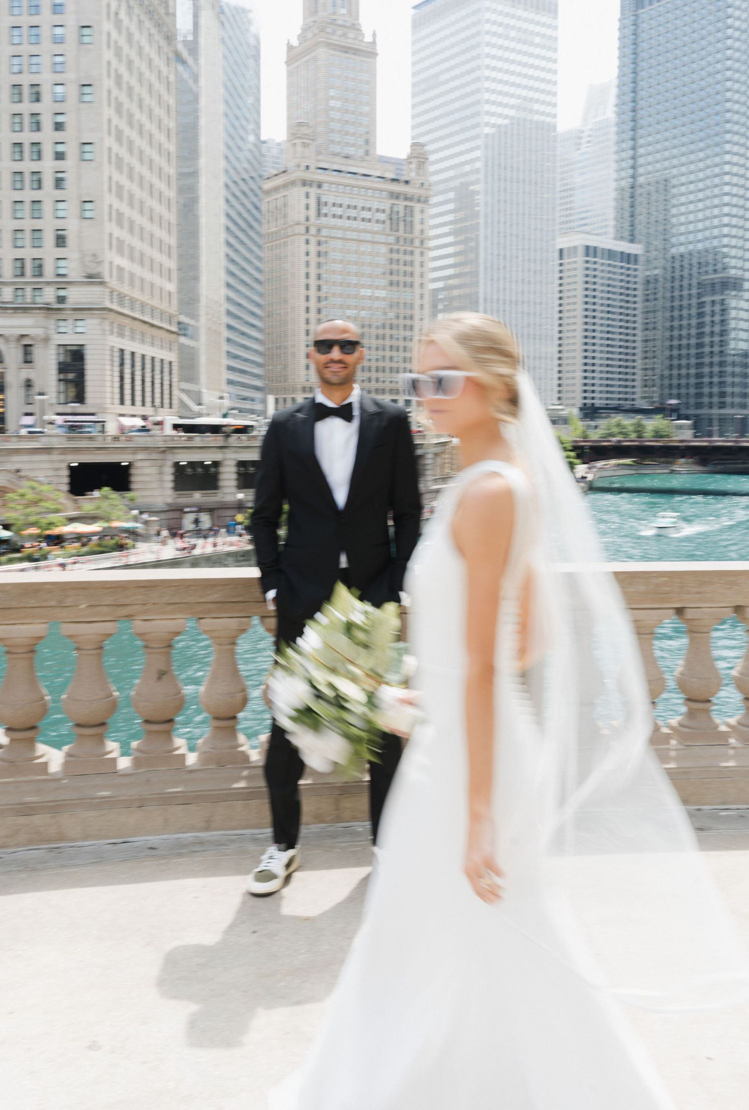 Bride and groom in sunglasses pose on a Chicago Riverwalk bridge, with city skyline and river in the background.