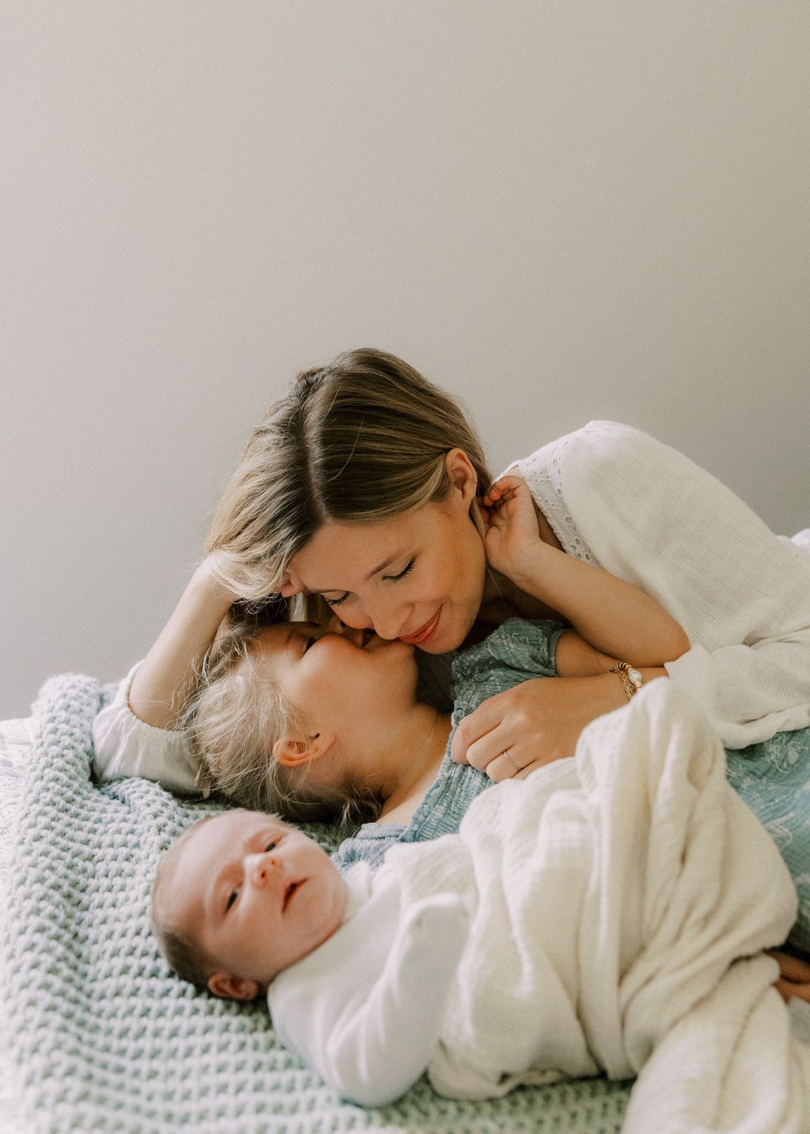 Mother tenderly kissing older child while newborn baby sleeps on soft blanket in cozy family moment.