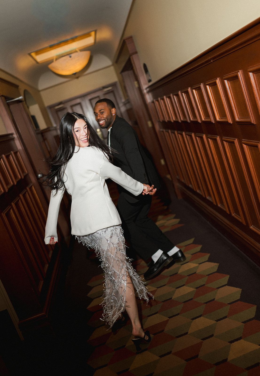 Couple holding hands walking down an elegant wood-paneled hallway, woman in white feather-hem mini dress.
