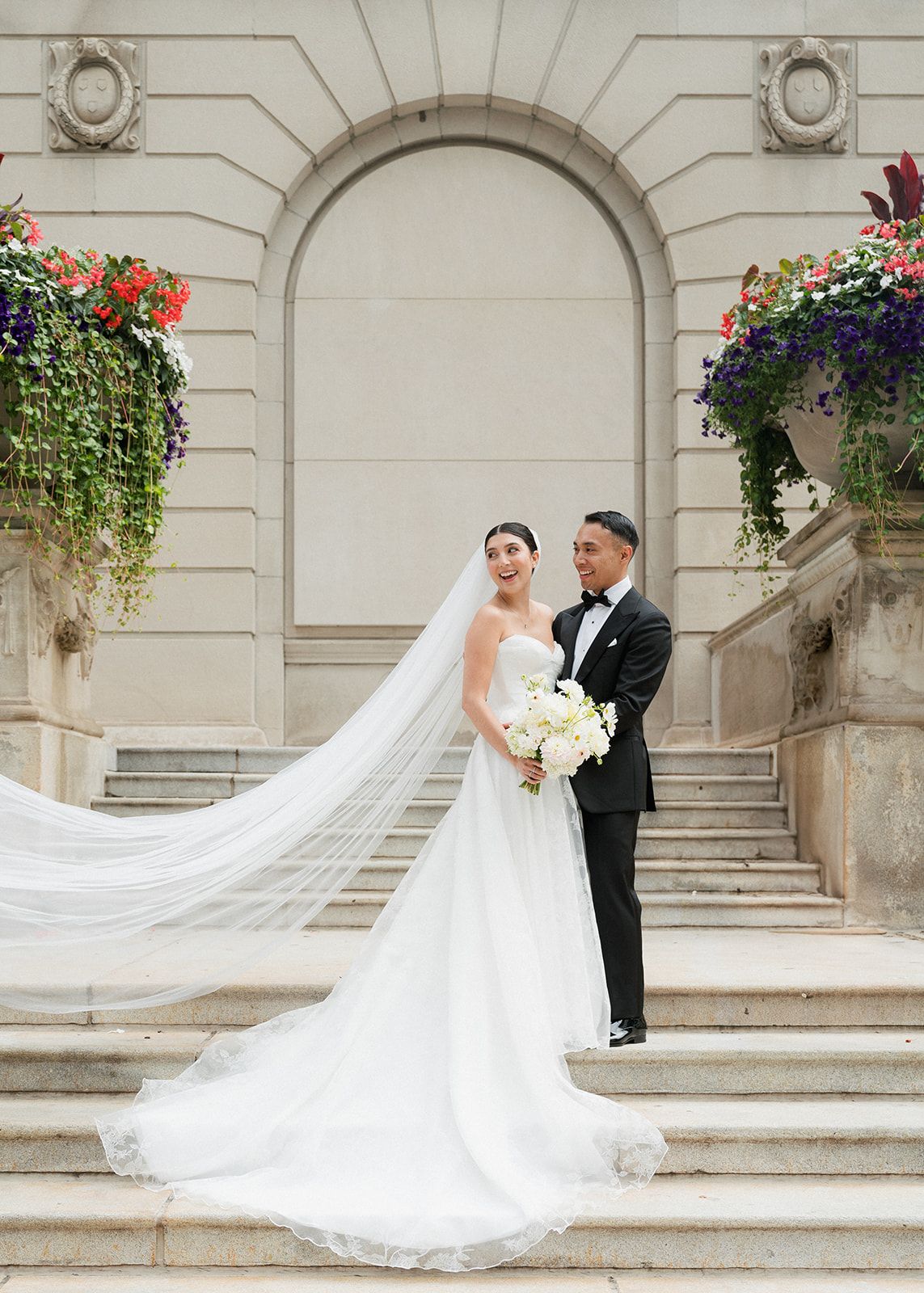 Bride and groom pose on grand steps with flowing cathedral veil and colorful floral arrangements.