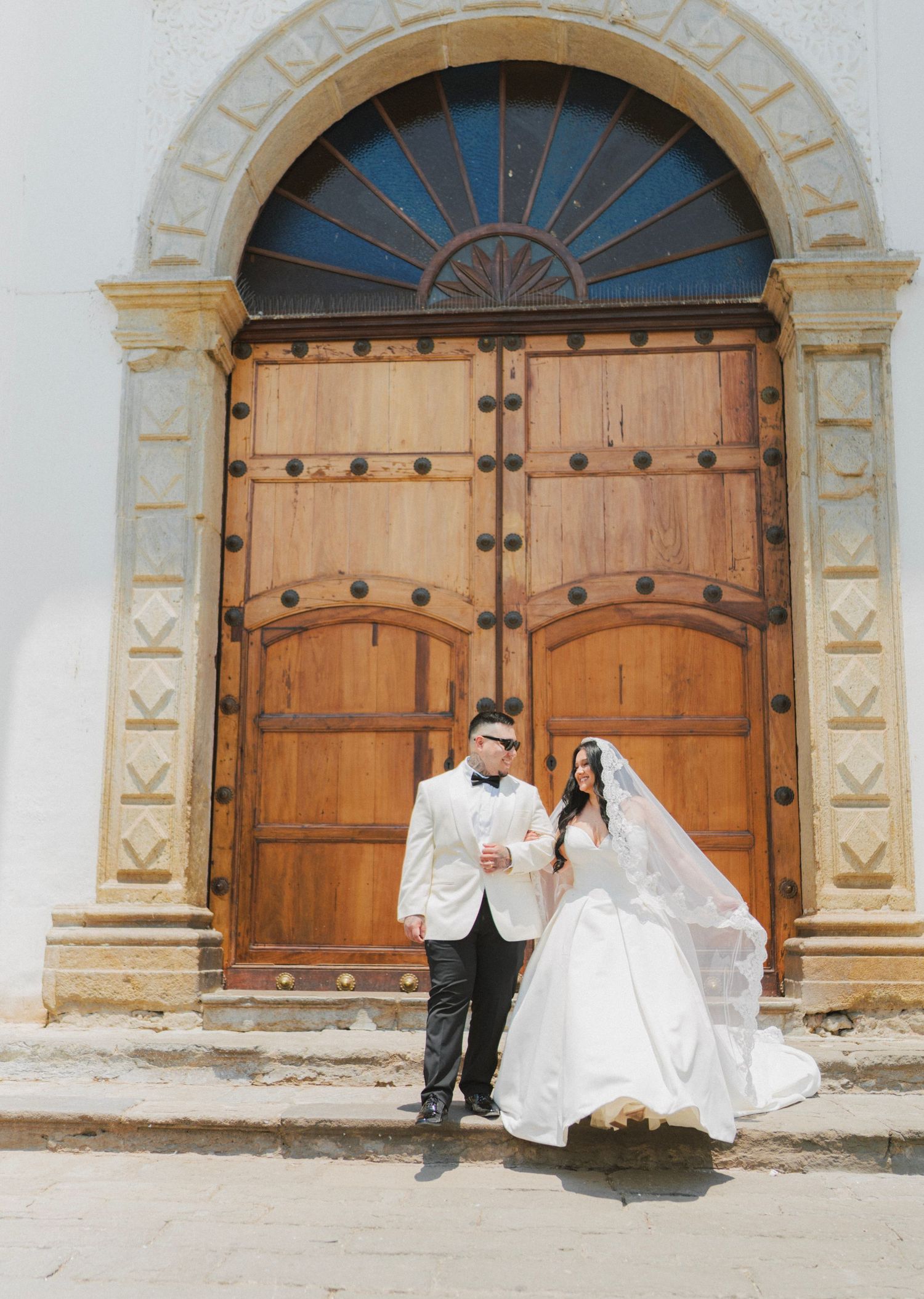 Bride and groom posing in front of ornate wooden church doors, bride in flowing white gown with veil, groom in white tuxedo.