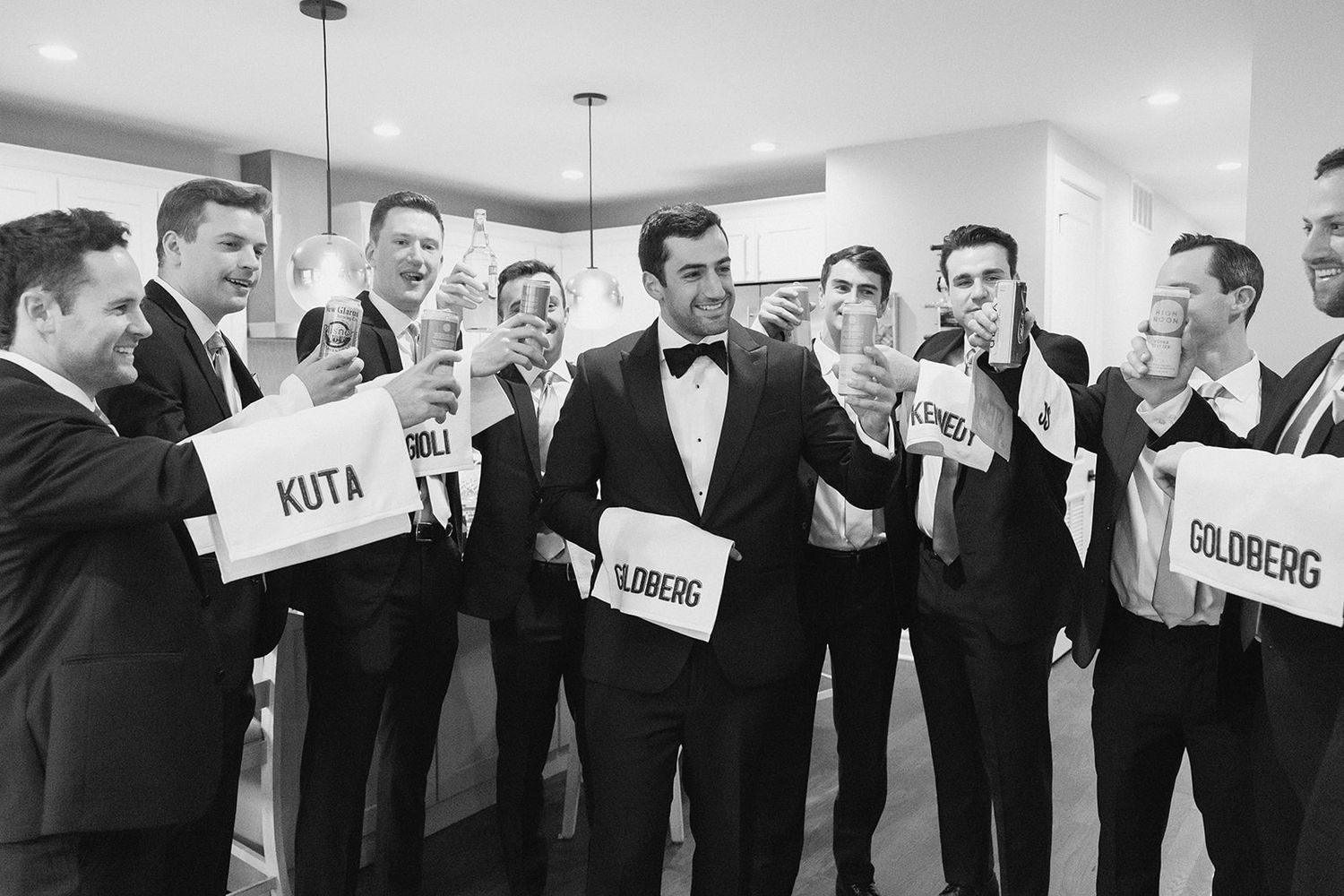 Groom surrounded by groomsmen in tuxedos holding name cards and celebrating in black and white photo.