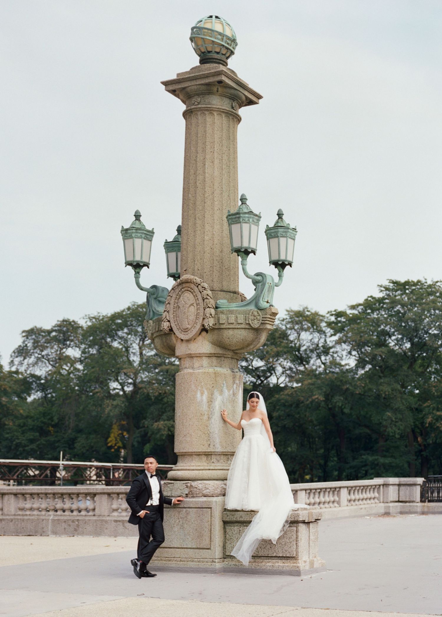 Bride and groom pose beside ornate stone column with lanterns at a grand outdoor venue.