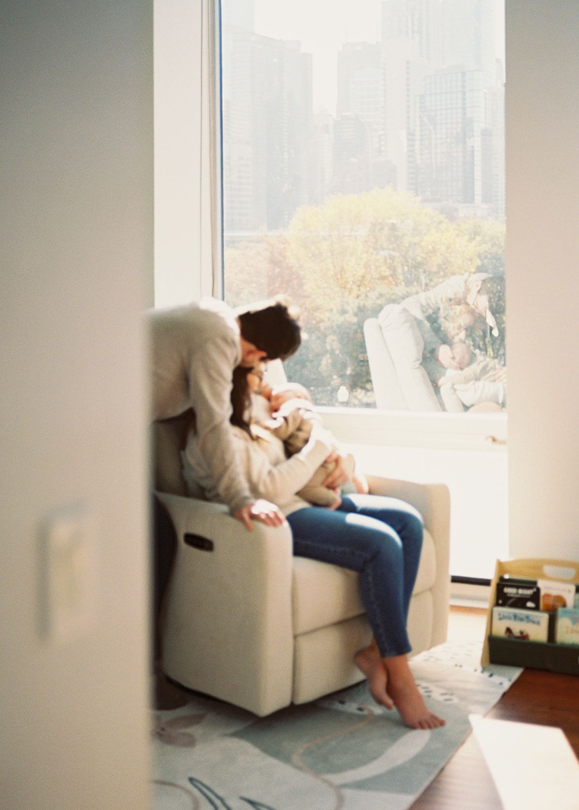 Mother sitting in a cozy chair by a large window, tenderly holding her newborn baby.
