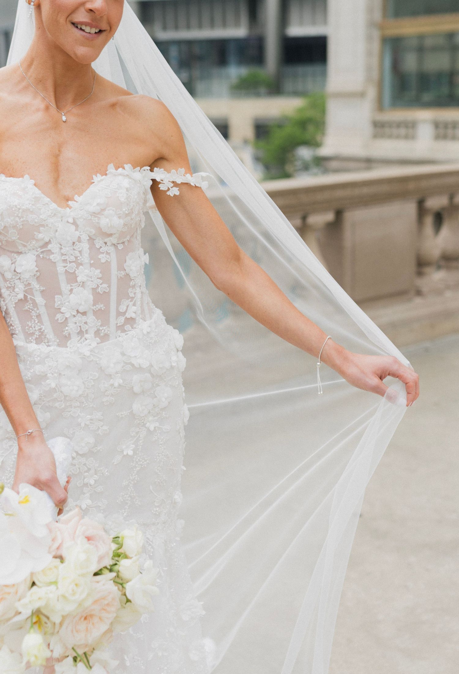 Bride in a floral lace off-shoulder wedding gown holding a white bouquet and flowing veil outdoors.