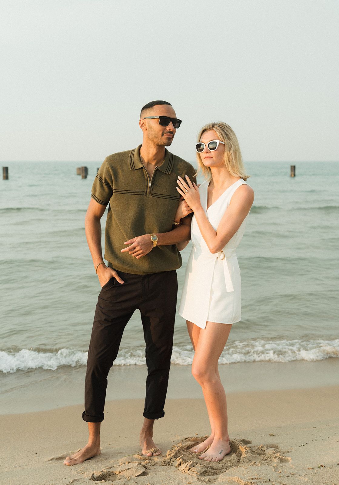 Couple in sunglasses posing on a sandy beach, man in olive polo and black pants, woman in white romper, ocean backdrop.