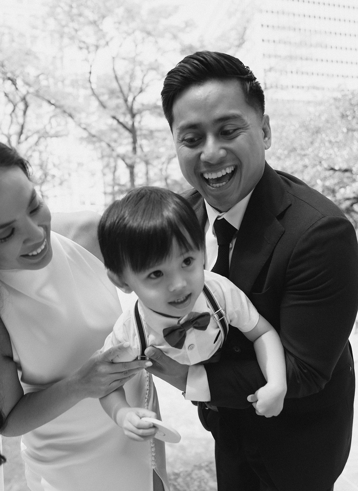 Black and white photo of a joyful family with a young boy in a bow tie outdoors.