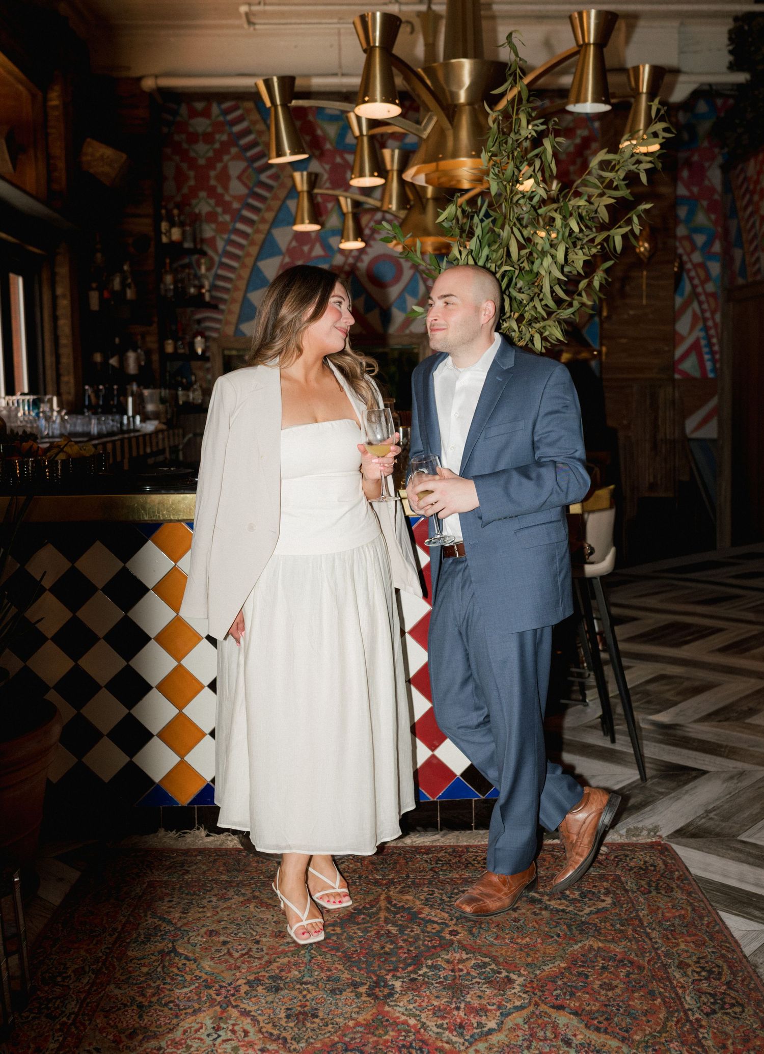 Couple in elegant attire holding drinks at a stylish bar with colorful mosaic tiles and gold pendant lights.