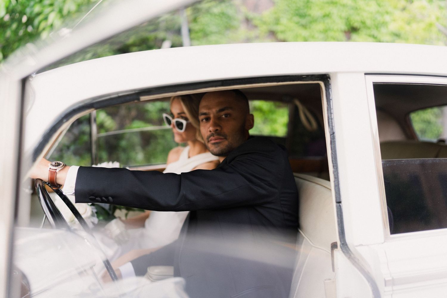 Groom in black suit sits at wheel of vintage white car with bride in sunglasses behind him.