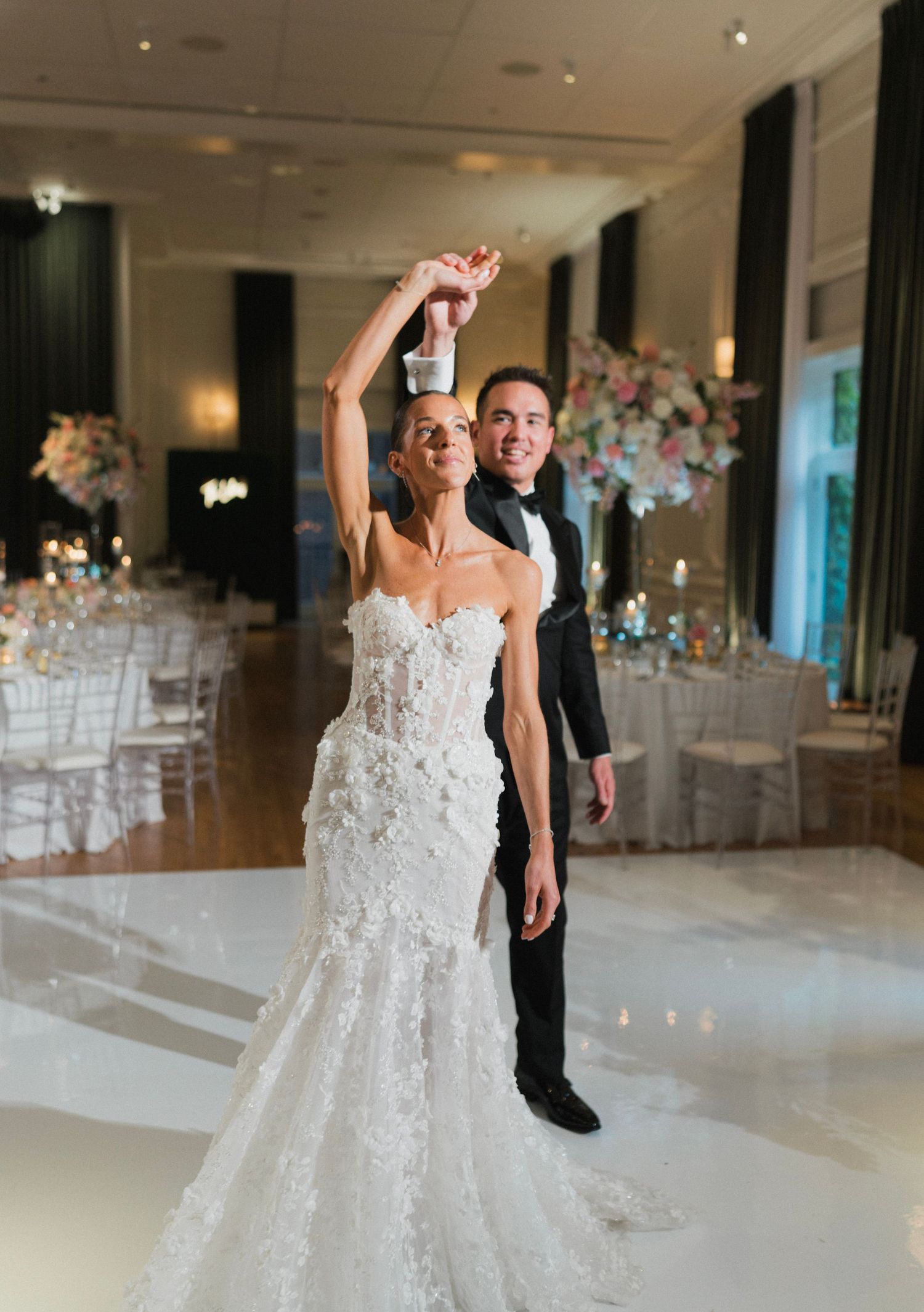 Bride in floral mermaid gown twirls on dance floor as groom in tuxedo spins her at elegant wedding reception.