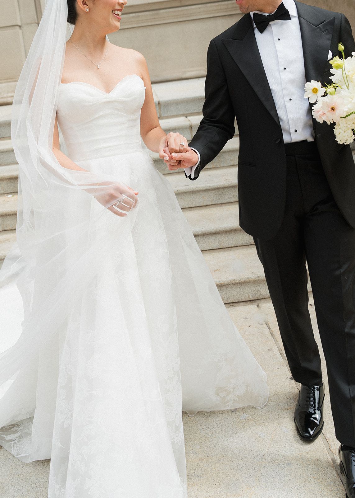 Bride in strapless sweetheart ballgown with cathedral veil holds hands with groom in black tuxedo on stone steps.