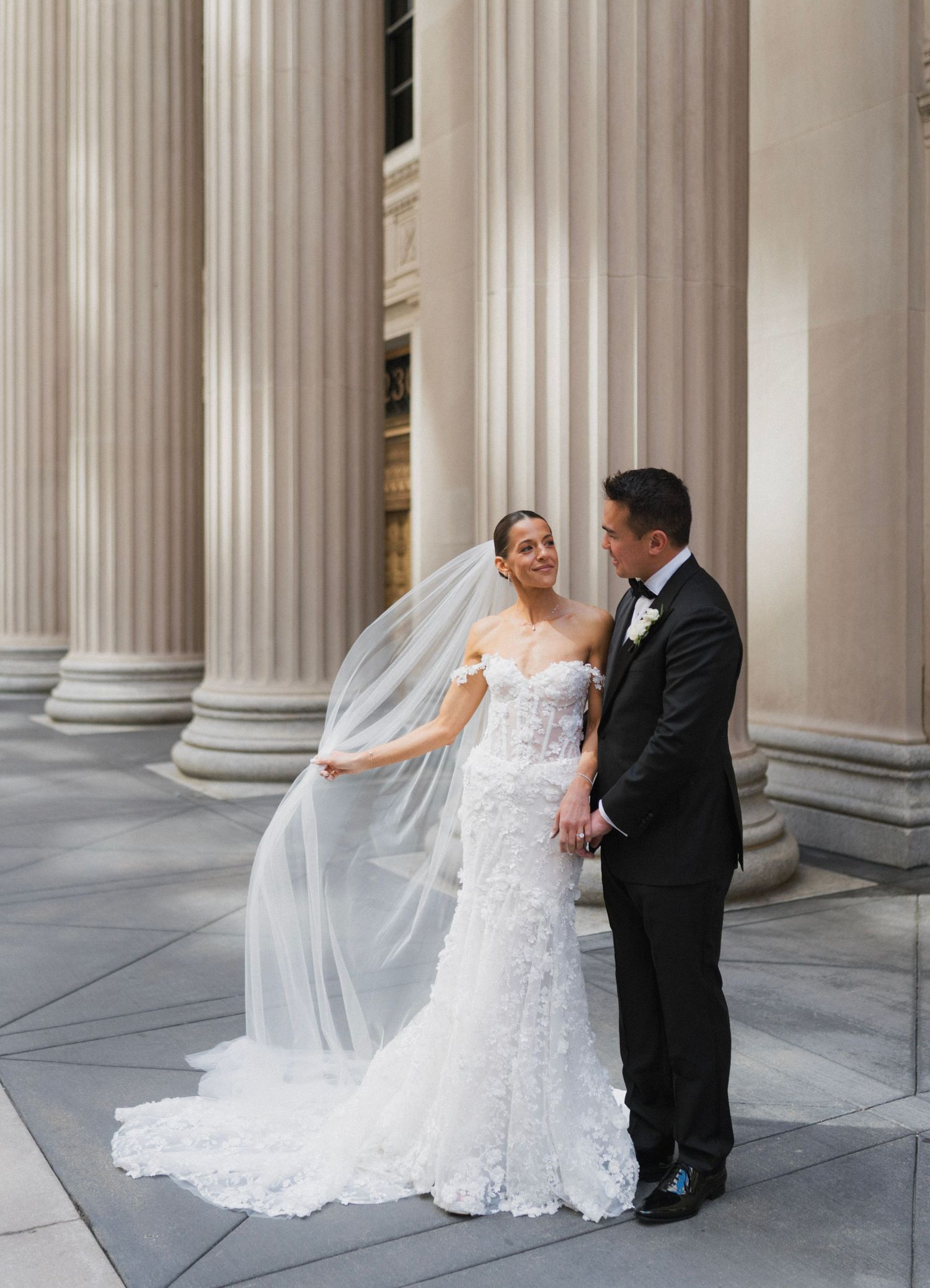 Bride in lace gown with flowing veil stands with groom in tuxedo before grand stone columns.