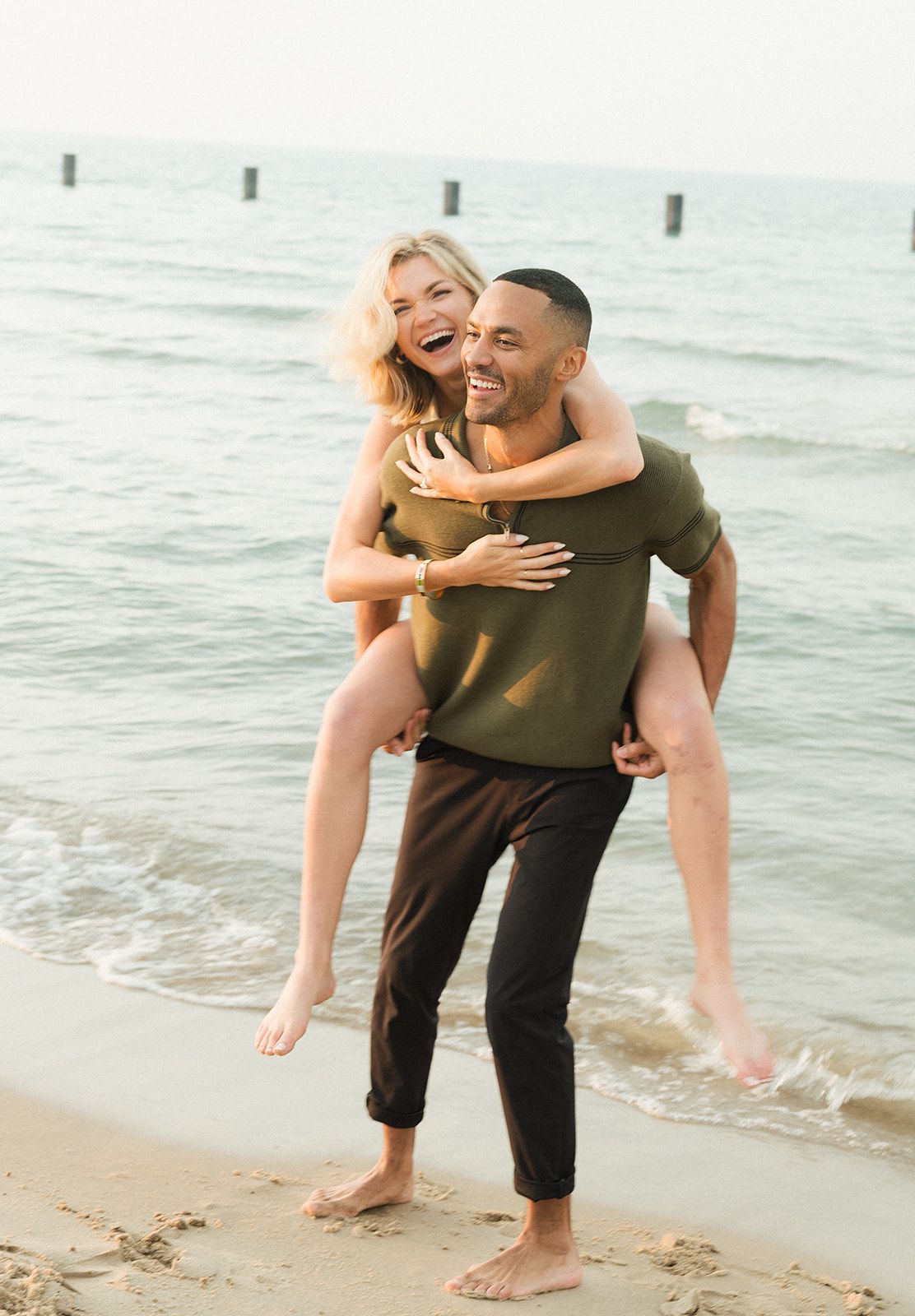 Laughing couple enjoying a piggyback ride on a sandy beach near the ocean waves.